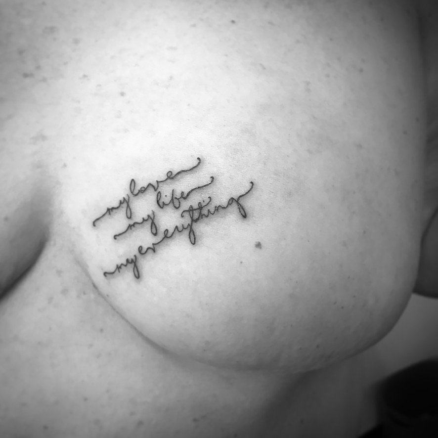 Close-up of a person’s breast with a tattoo in cursive writing that says, "my love my life my everything".