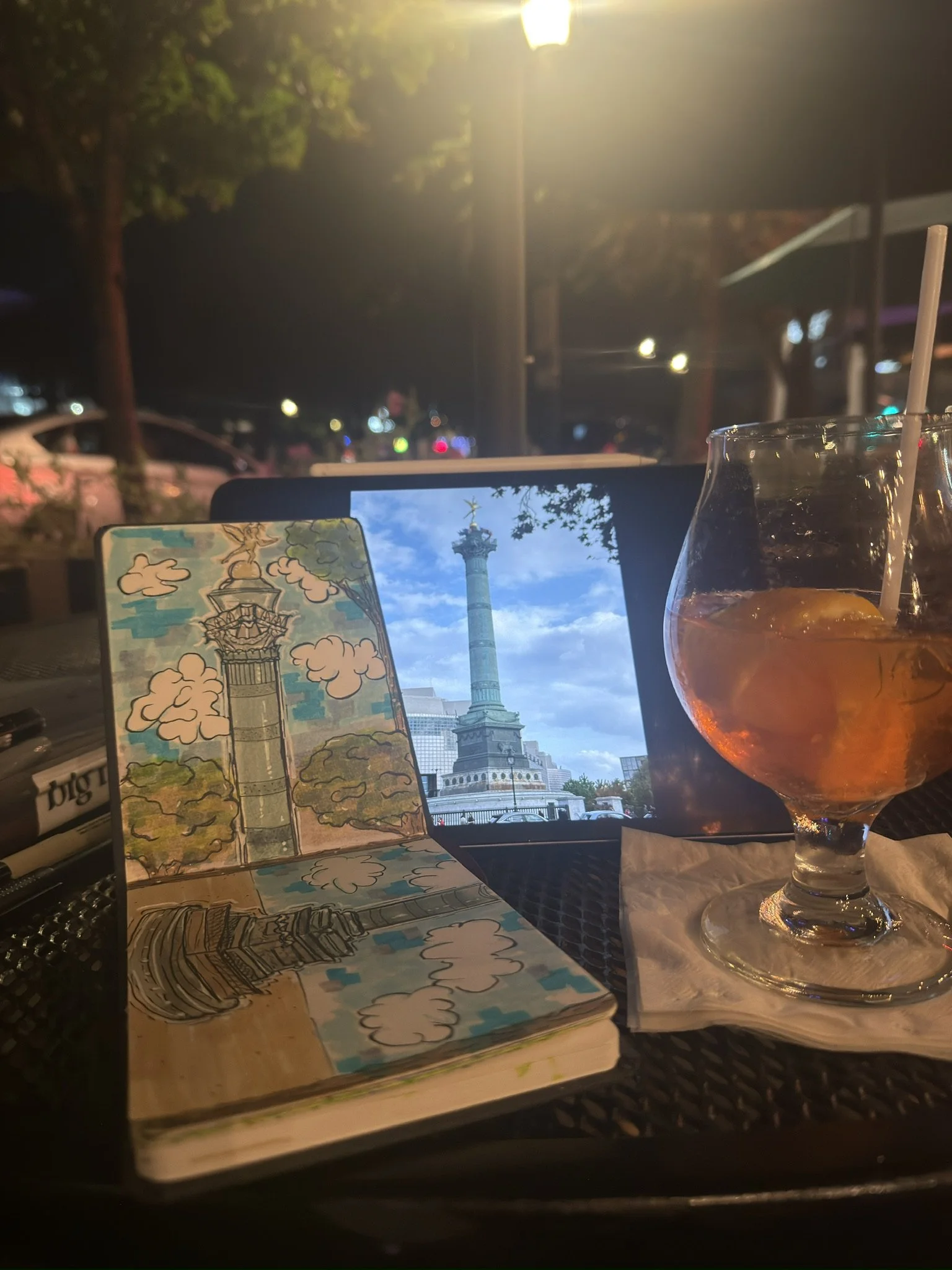 An outdoor table at night with a tablet displaying a photo of a monument, a colorful sketch of a similar monument, a cocktail drink with ice and lemon, and a napkin, with streetlights and trees in the background.