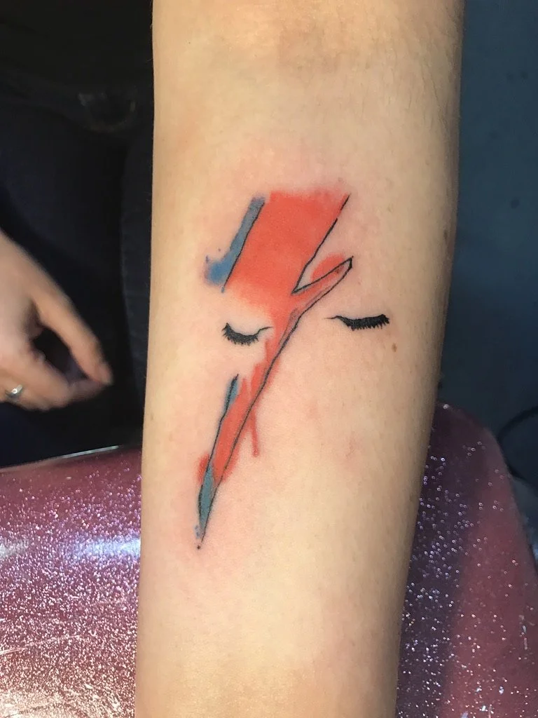 Tattoo of a stylized Aladdin Sane David Bowie face with closed eyes, featuring a bold red and black lightning bolt running through it on a person's forearm.