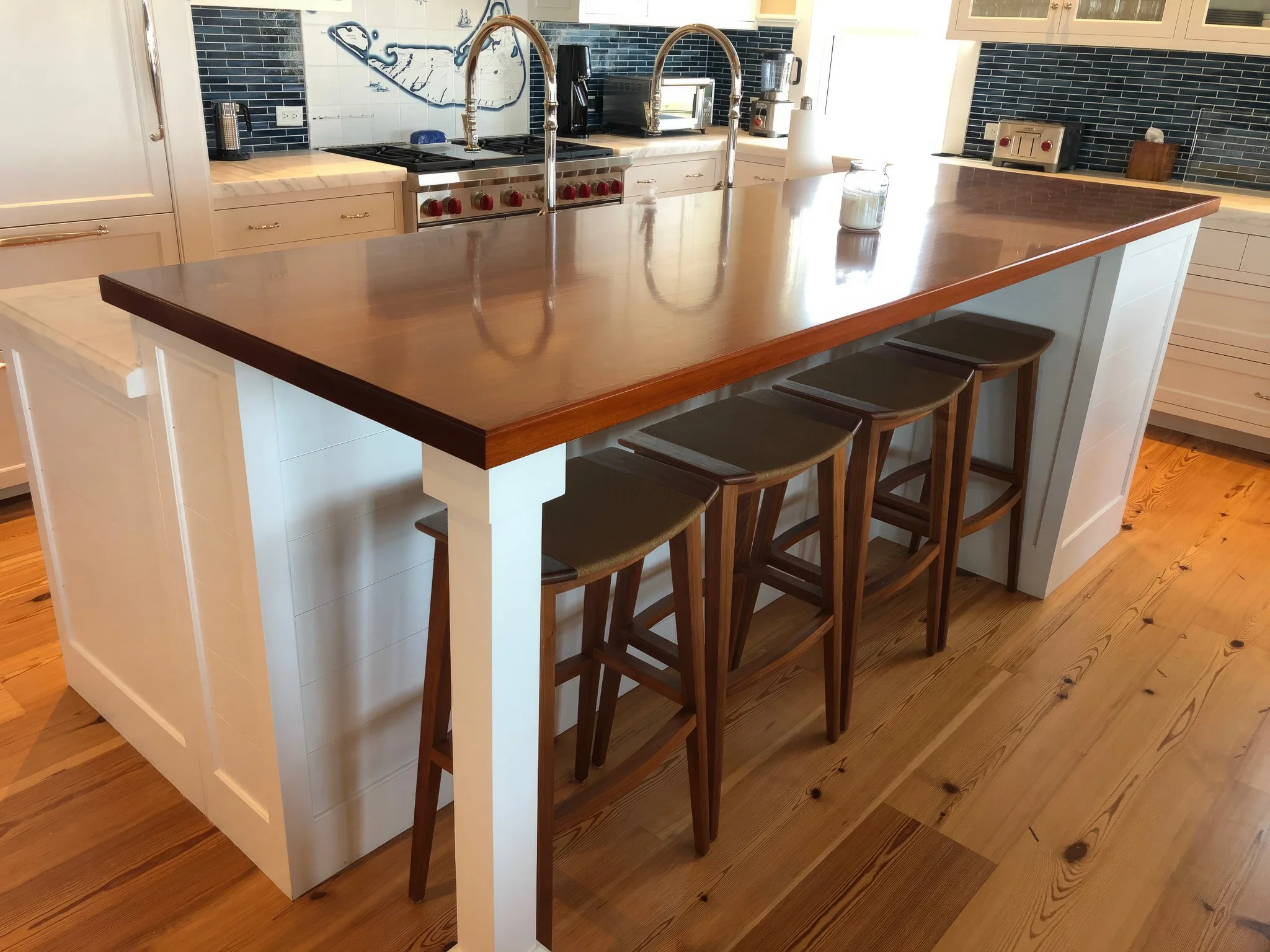 A modern kitchen island with a wooden countertop and three bar stools, with a stove, sink, and appliances in the background, costom kitchen, Boston custom carpentry, Nantucket custom kitchen, West Newbury based custom carpentry