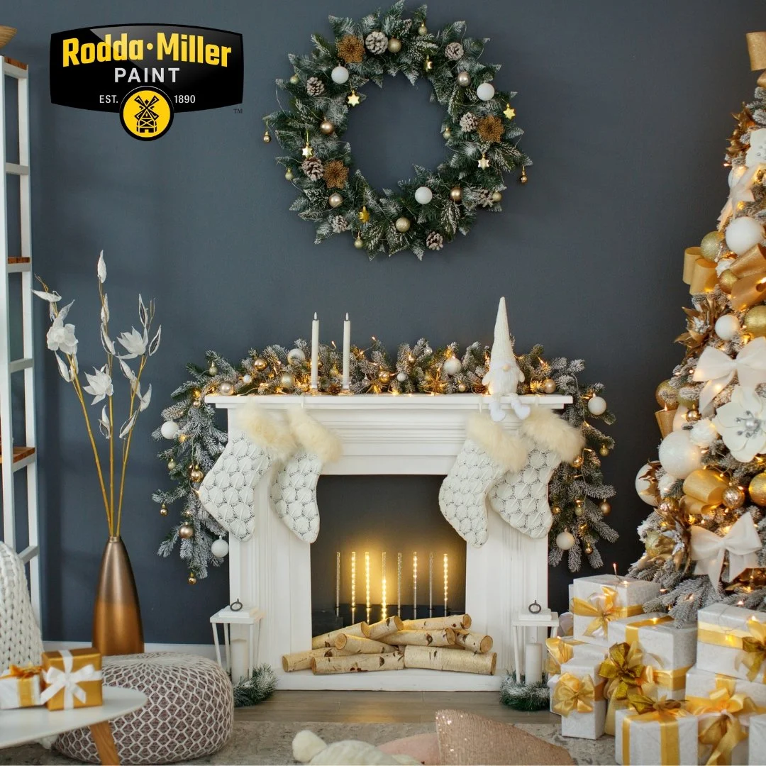 A festive holiday living room with a decorated Christmas tree, a white fireplace mantle with stockings, candles, and ornaments, a wreath on the wall, wrapped gifts with gold ribbons, a tall gold vase with decorative branches, and cozy seating.