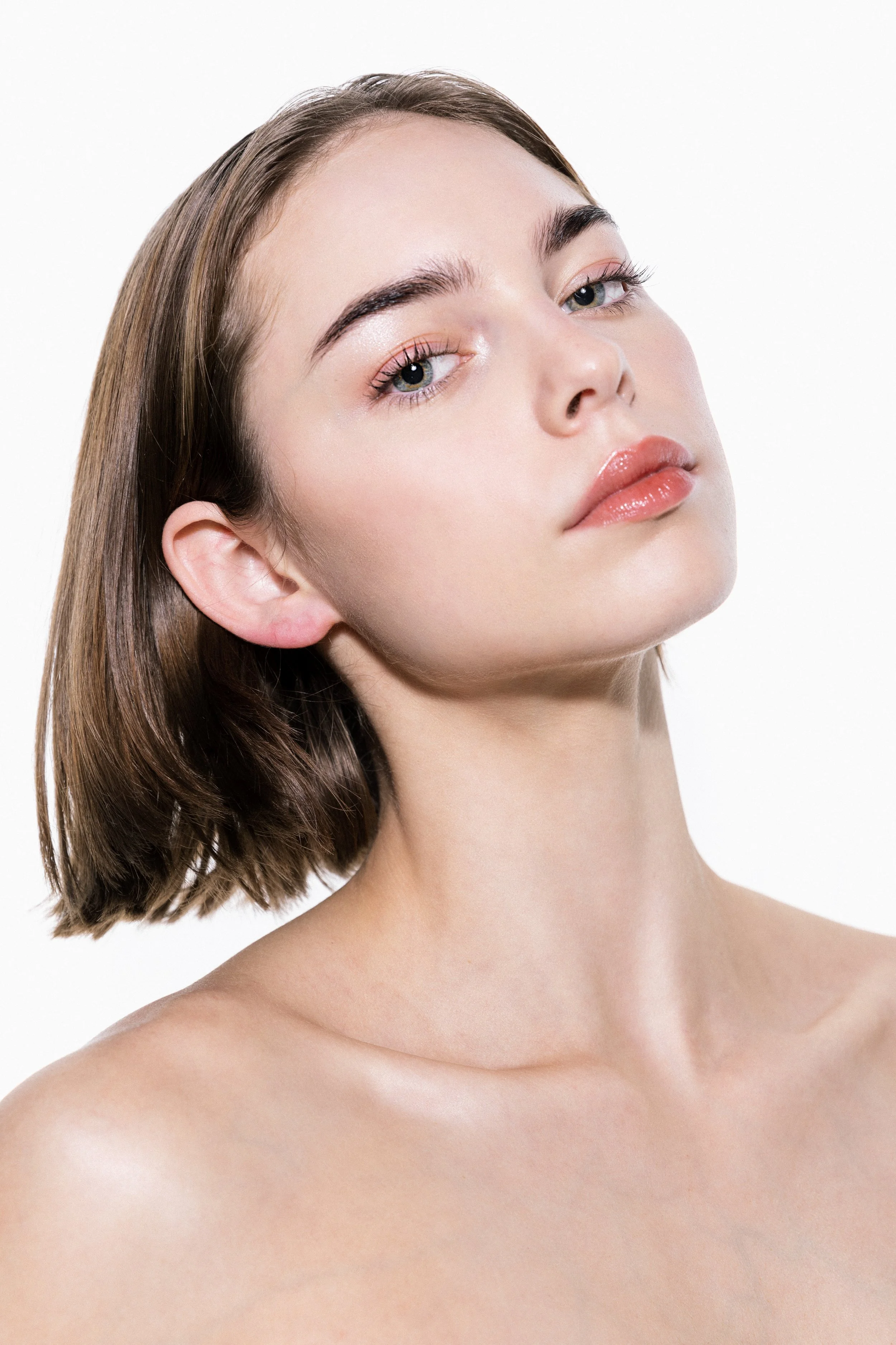 The model's pouty lips and bright eyes take centerstage against a subtle, dewy base. A peachy-pink gloss mirrors a sweep of coral shadow on her lids. The look comes together with a light layer of mascara, brow gel, and inner corner highlight, creatin