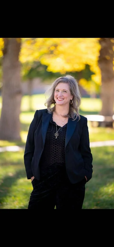 Woman in a black blazer and black top smiling outdoors in a park with trees and yellow autumn leaves.
