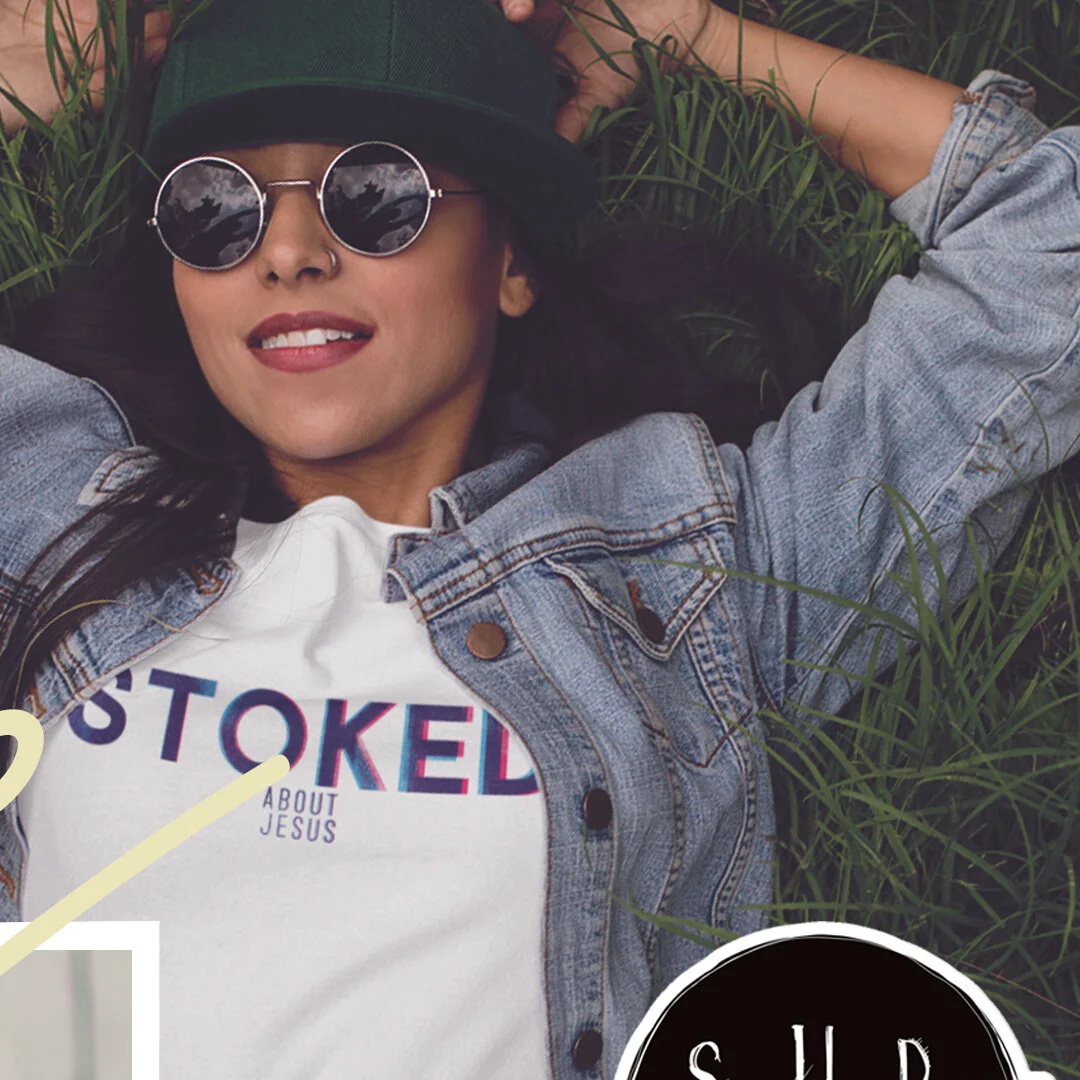 Woman lying on grass wearing sunglasses, a green hat, a denim jacket, and a white T-shirt with text about Jesus.