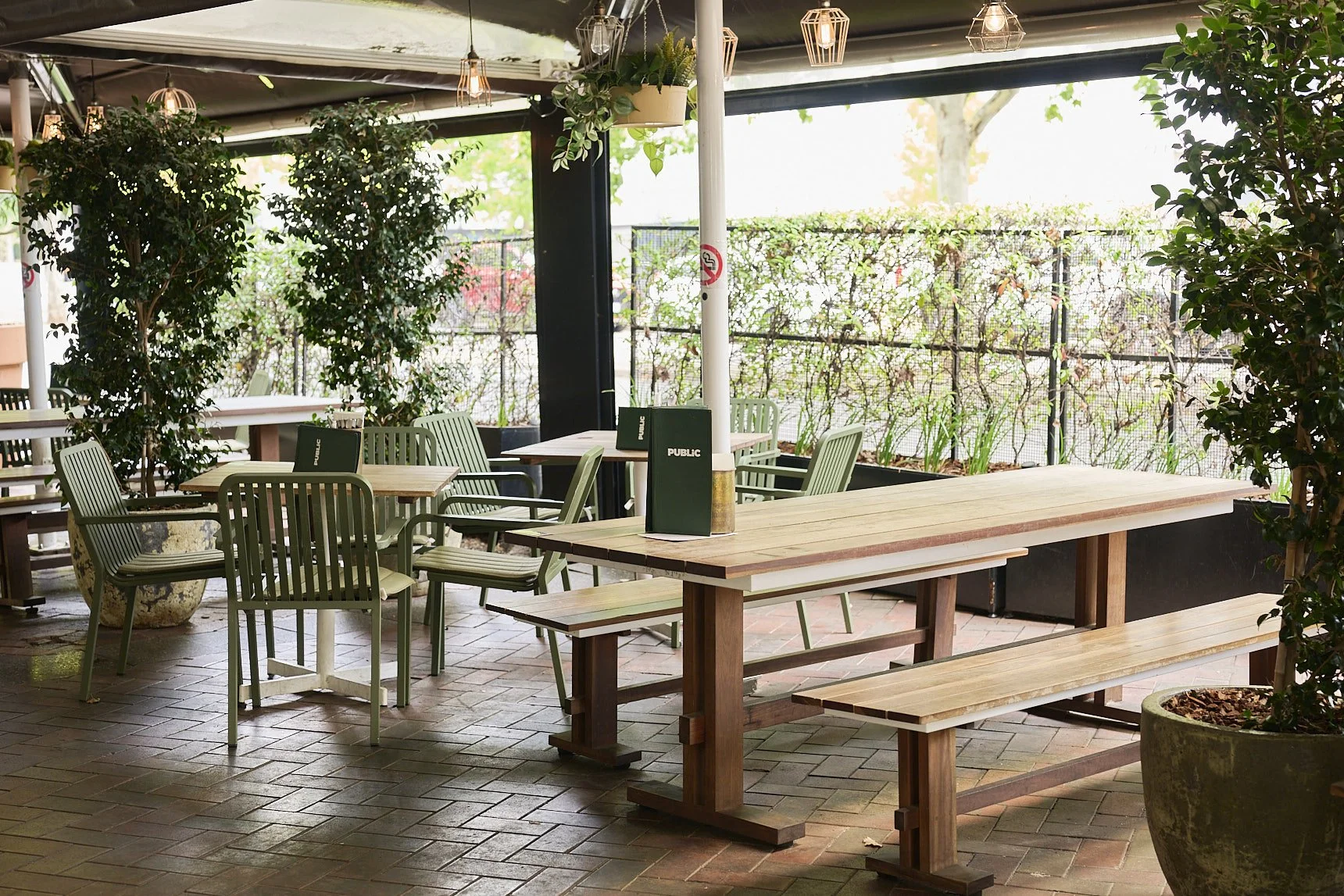 Empty outdoor beer garden with wooden tables, white chairs, potted plants, and large windows with curtains allowing sunlight to fill the space.