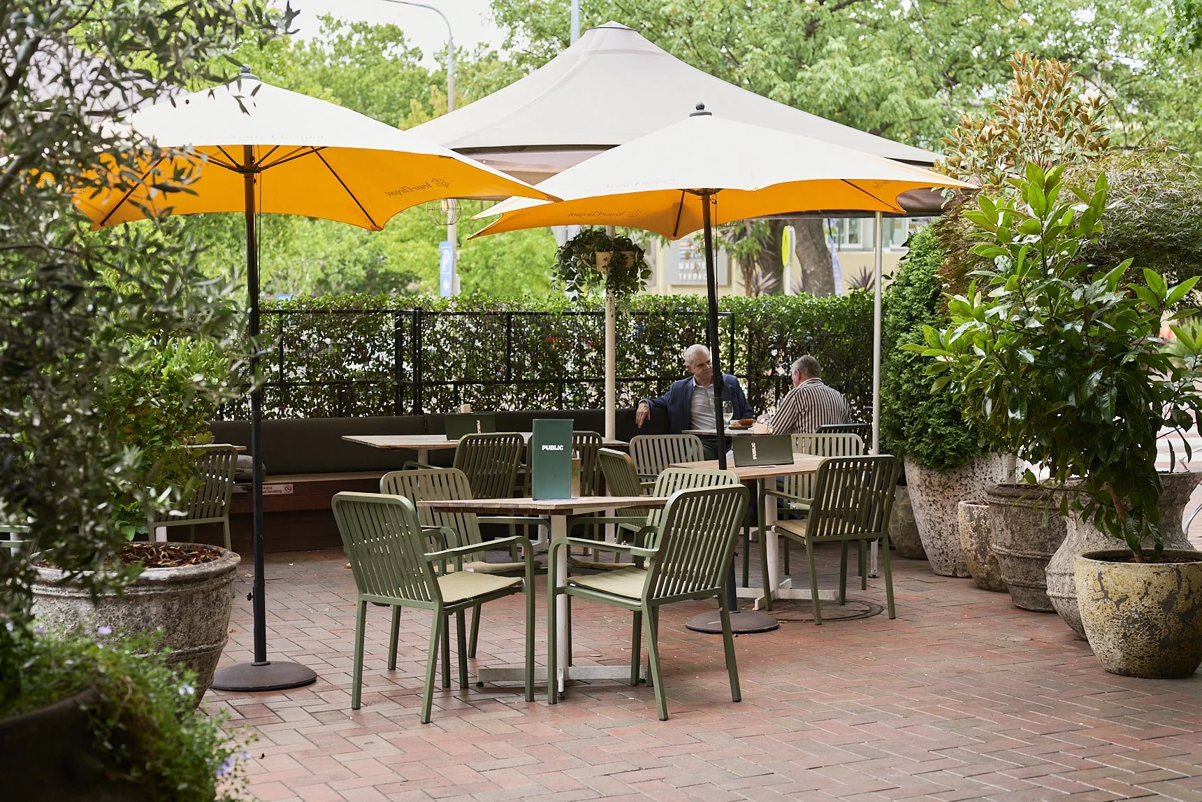 Outdoor courtyard with green chairs around wooden tables, surrounded by potted plants and heaters, under a gazebo with hanging lights