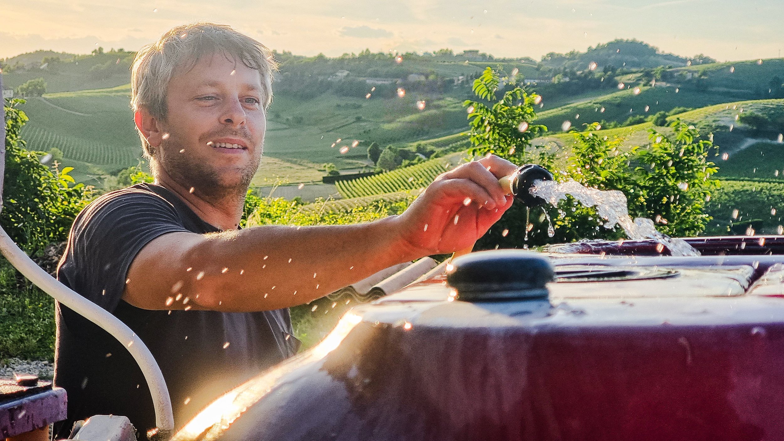 A man is pouring water from a hose onto a red surface outdoors, with green hills and trees in the background during sunset.