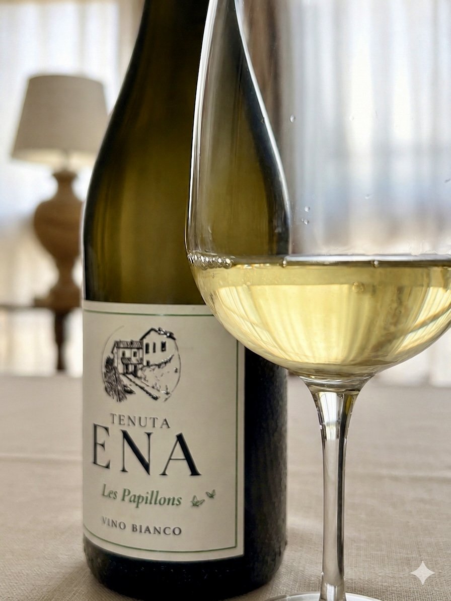 A bottle of Tenuta Ena white wine labeled "Les Papillons" next to a filled wine glass, on a beige tablecloth, with a blurred background of a lamp and curtain.