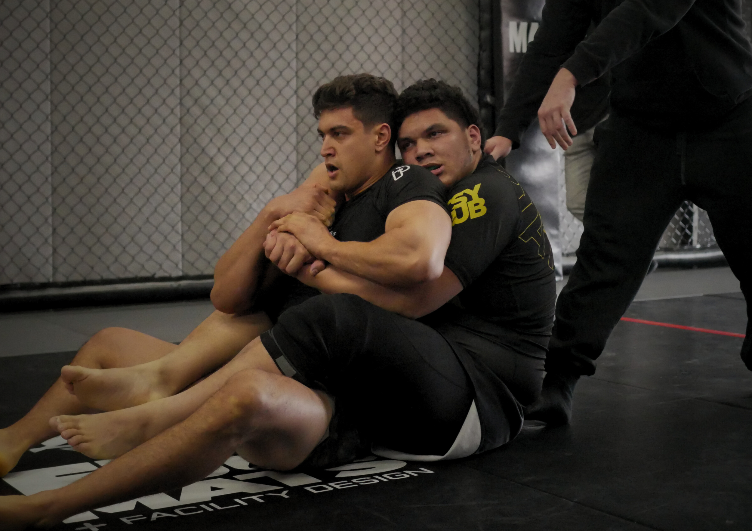 Two male athletes engage in a grappling match inside a fencing ring, with one on top of the other. The athlete on top has a focused expression, while the other appears to be resisting. They are both wearing black athletic gear, and a referee or coach
