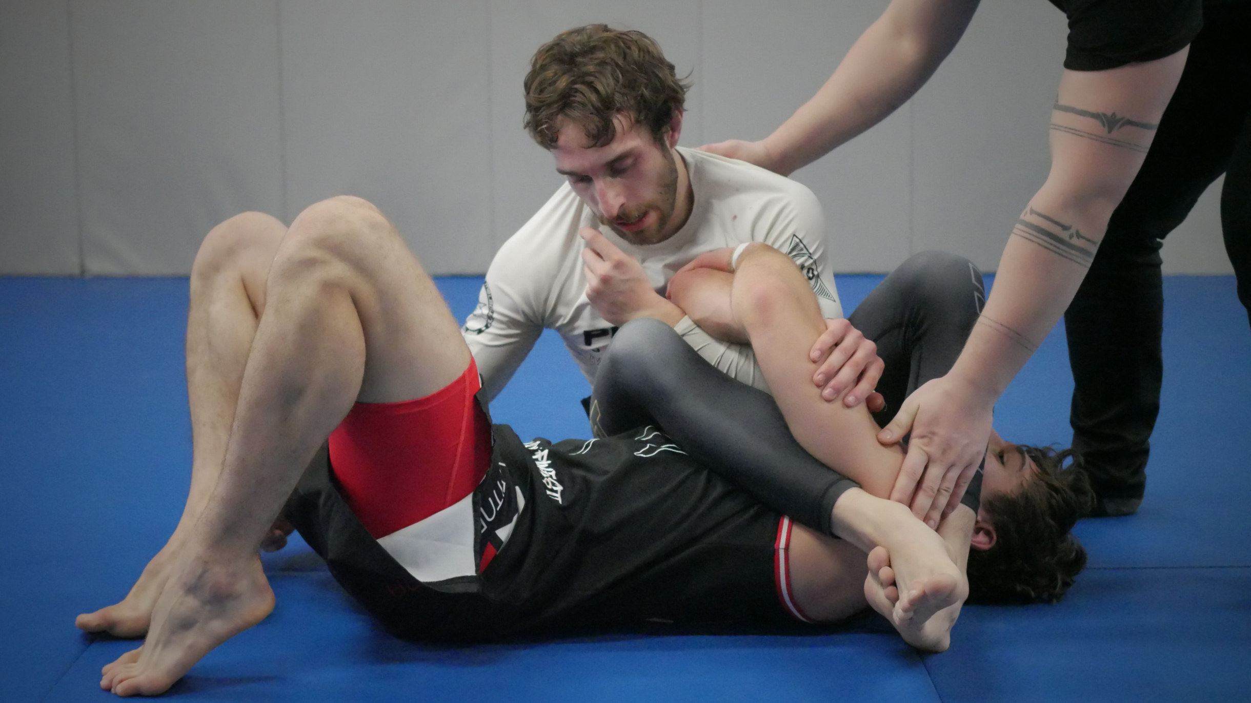 Two male mixed martial arts fighters on the ground in a grappling position with a referee nearby, on a blue padded mat in a training room.