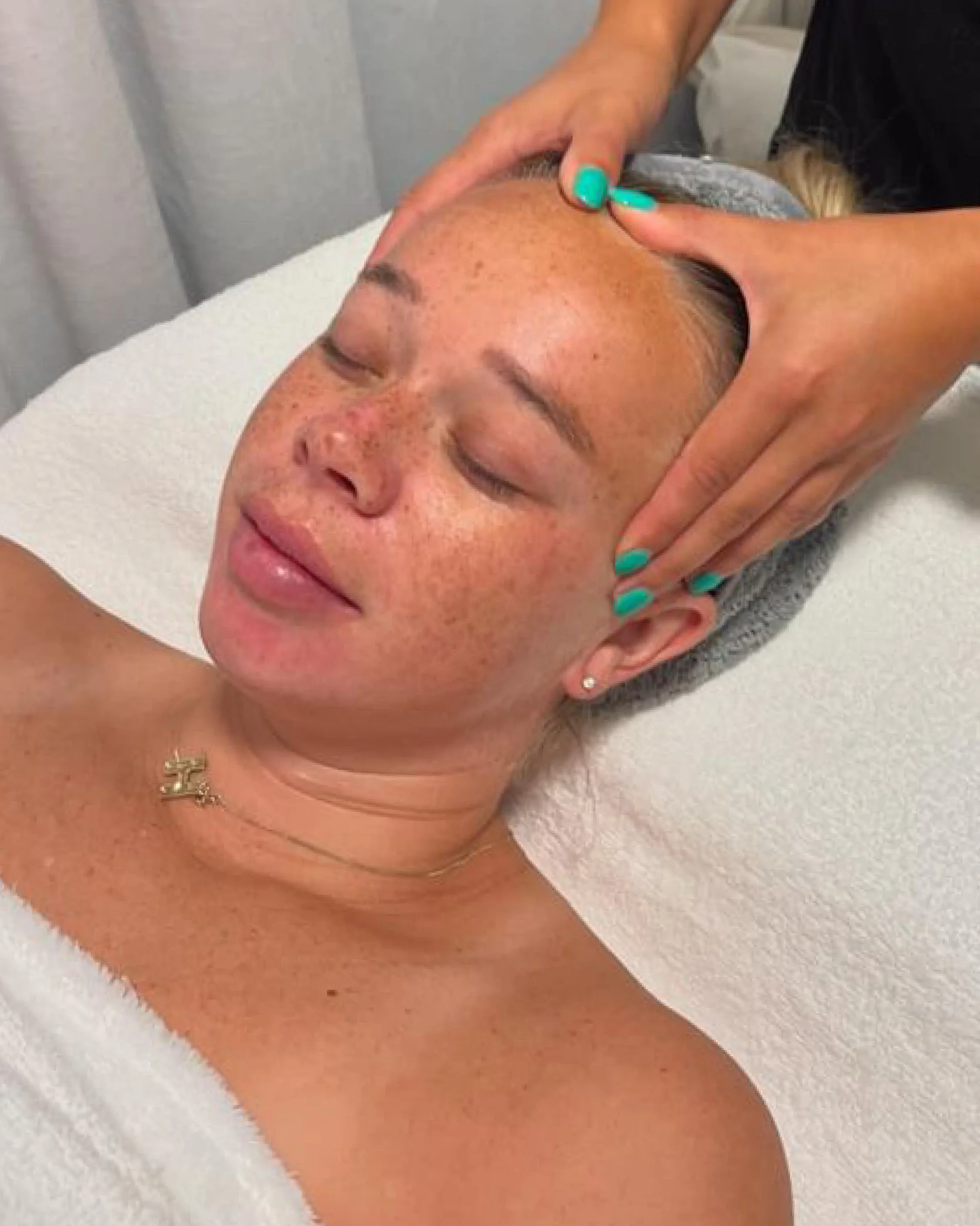 Person receiving a facial massage with eyes closed, lying on a white towel, while another person with teal nail polish applies pressure to their forehead.