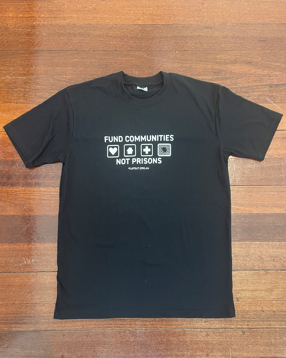 Black tshirt with white design on the chest. 4 squares show a heart, a home, a hospital cross and chain wire with a hole in it. Fund communities not prisons. flatout.org.au in small letters.