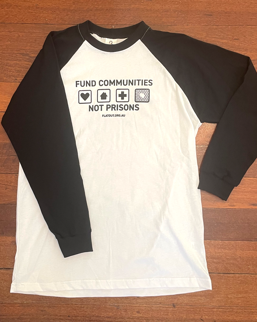 White tshirt with long black sleeves. Black design printed on the chest. 4 squares with heart, home, hospital and chain wire with a hole. Fund communities not prisons in small letters.