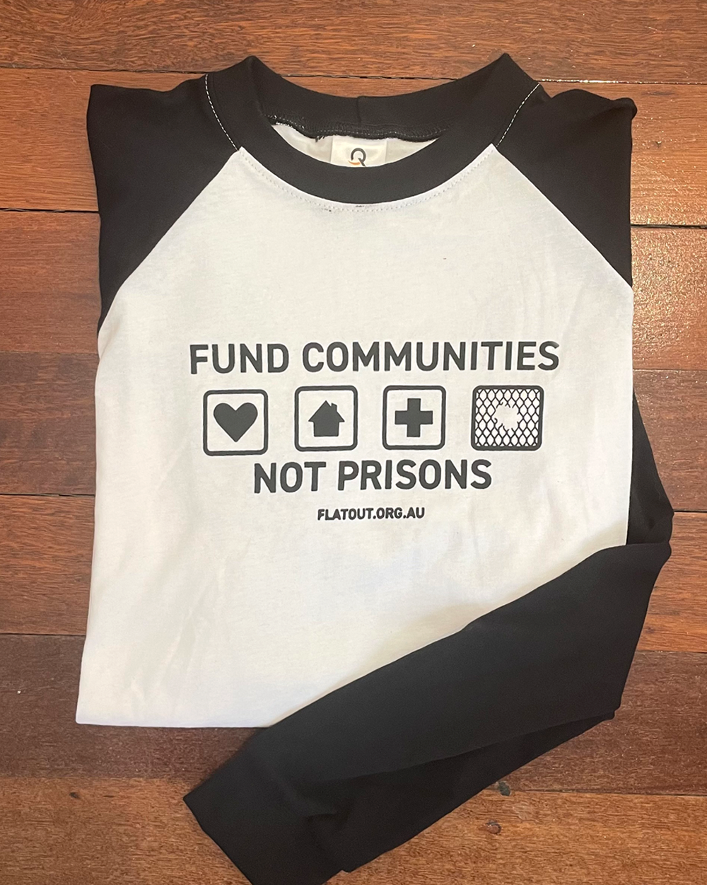 White tshirt with long black sleeves. Black design printed on the chest. 4 squares with heart, home, hospital and chain wire with a hole. Fund communities not prisons in small letters.