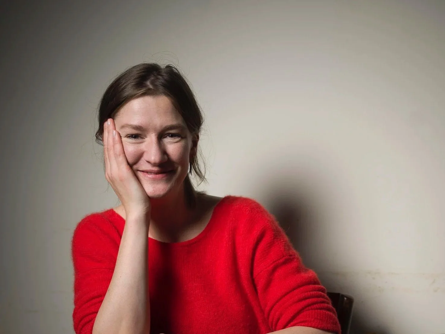 A woman with brown hair wearing a red sweater, smiling softly with her hand resting on her cheek, sitting against a plain light-colored wall.