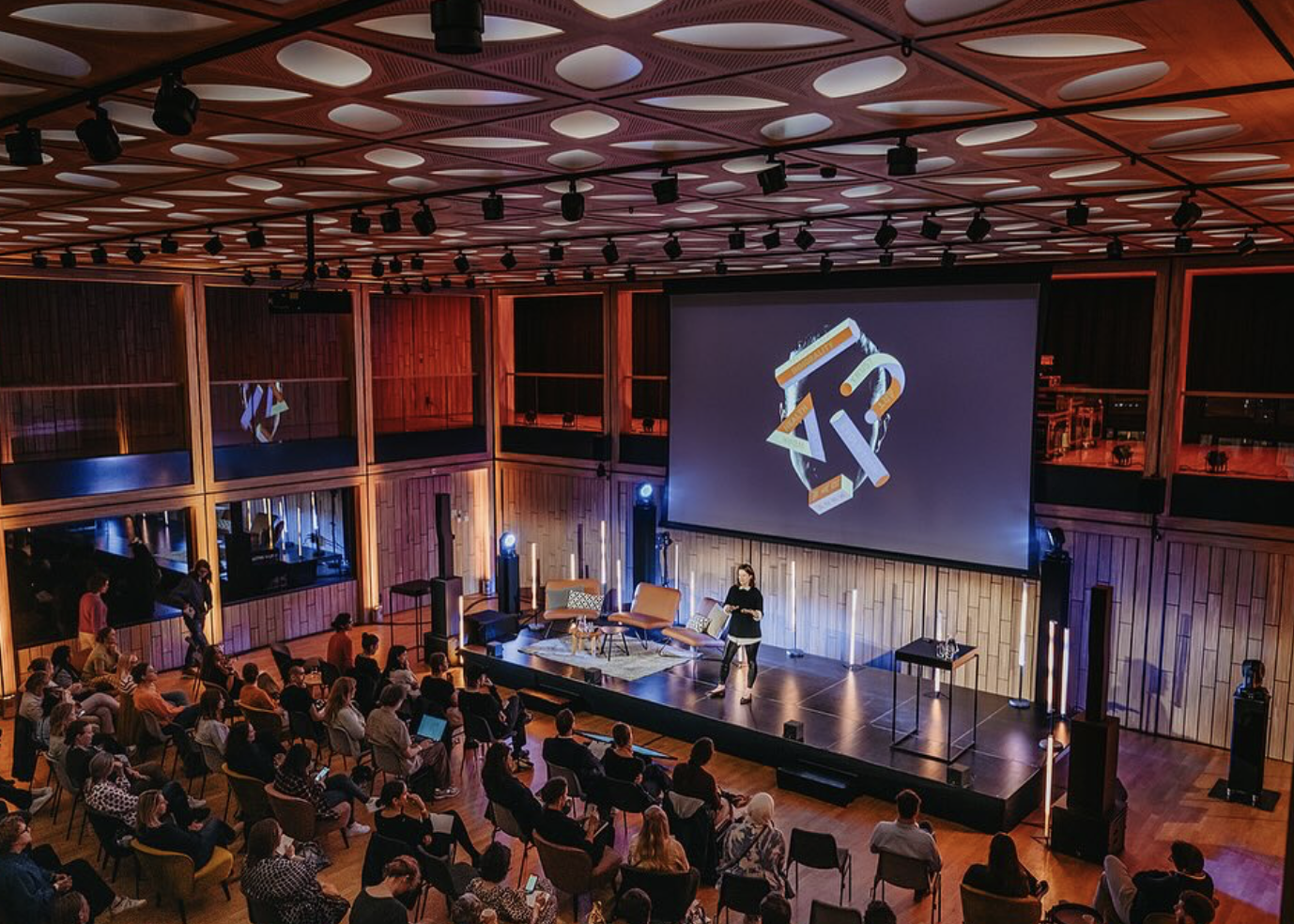 A speaker presents on a stage in a modern auditorium with an audience seated facing the stage. The stage has chairs, a small table, and a large projection screen displaying an abstract colorful 3D geometric design. The auditorium has wood-paneled walls, a patterned ceiling with circular lights, and glass-enclosed upper balcony areas.