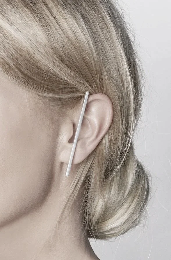 Una persona con el cabello rubio peinado hacia atrás, usando un pendiente largo y recto en la oreja. El fondo es blanco o gris claro.