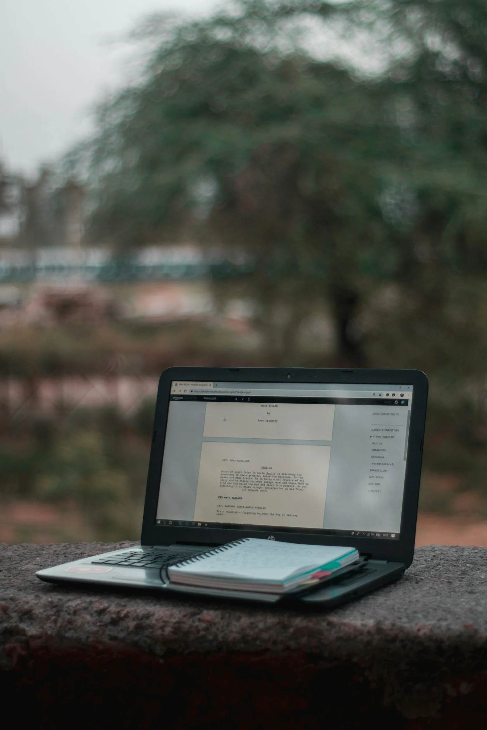 A laptop with a notebook on top, displaying a document on its screen, placed outdoors on a stone surface with blurred trees in the background.