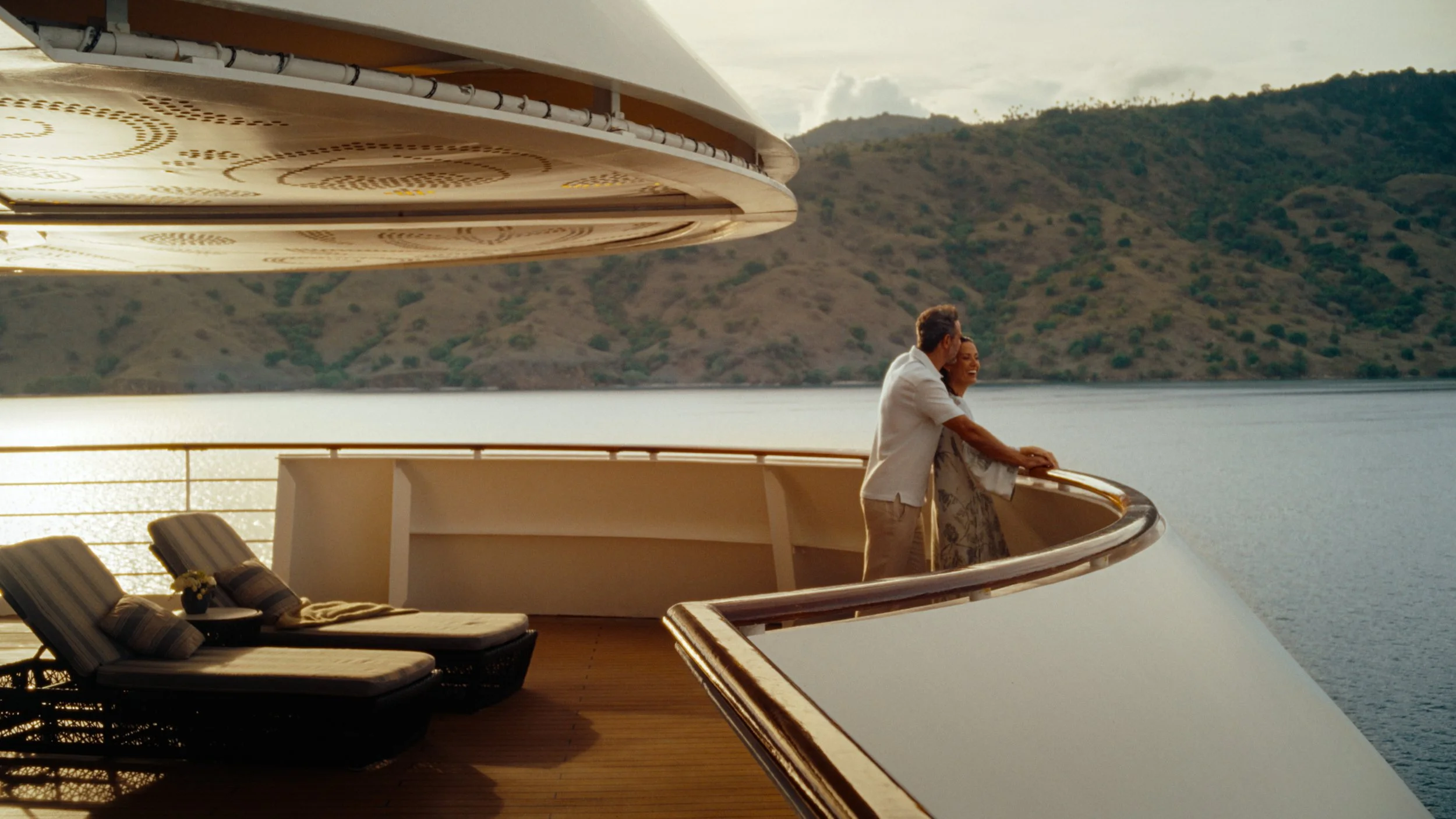 A couple standing on the deck of a yacht, looking out over the water with a scenic hillside in the background during sunset.