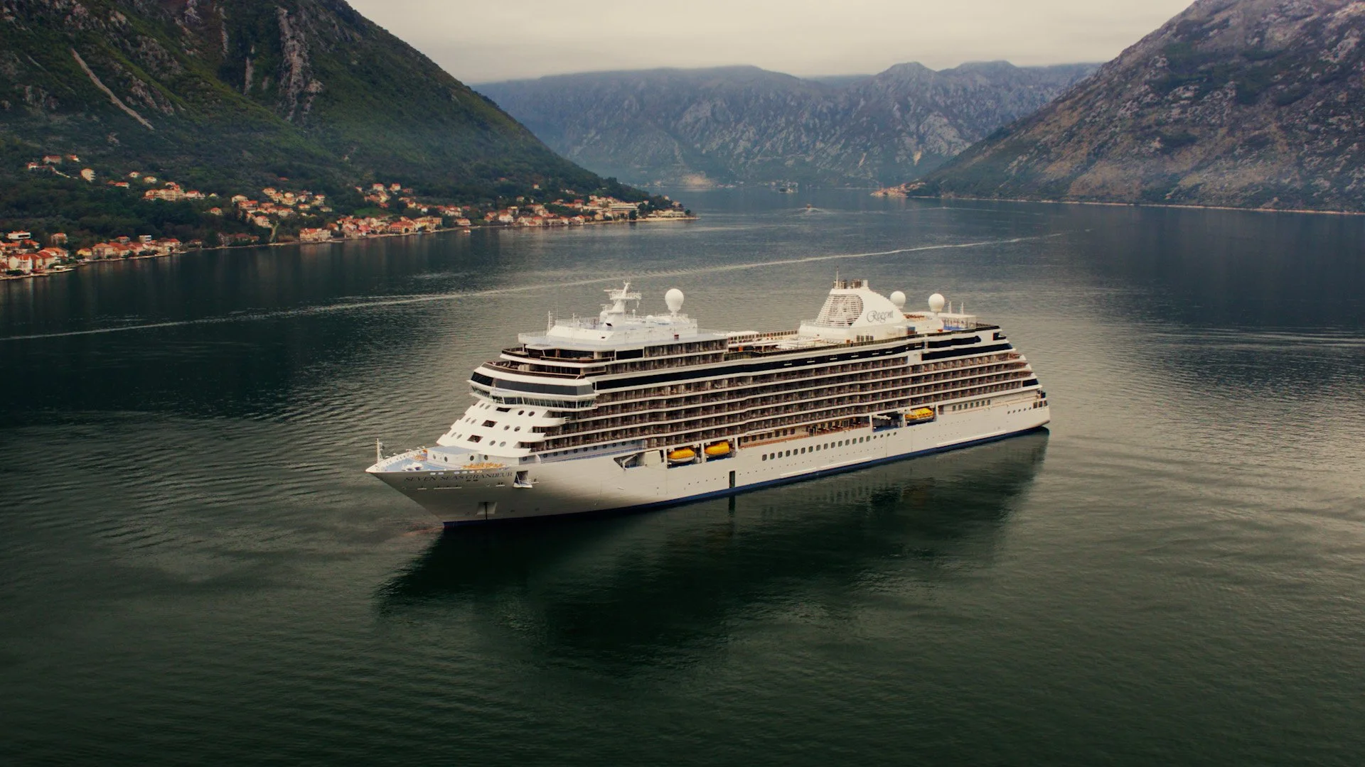 A large white cruise ship sailing through a fjord surrounded by green mountainous landscape with small houses along the shoreline.
