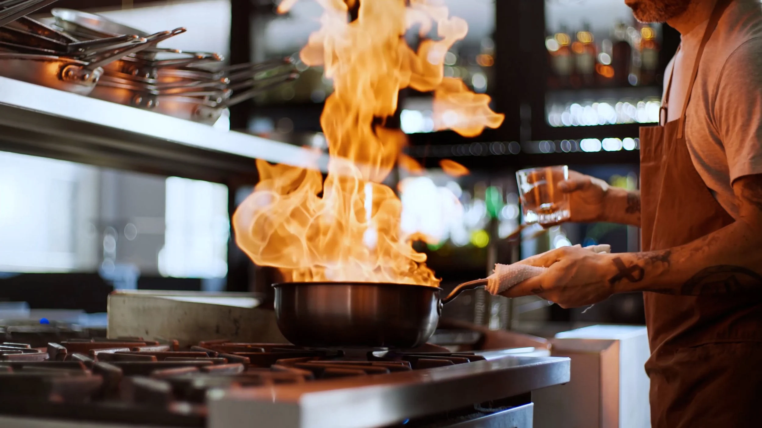 A chef at a restaurant kitchen holding a glass cup, with a pan on the stove emitting flames.