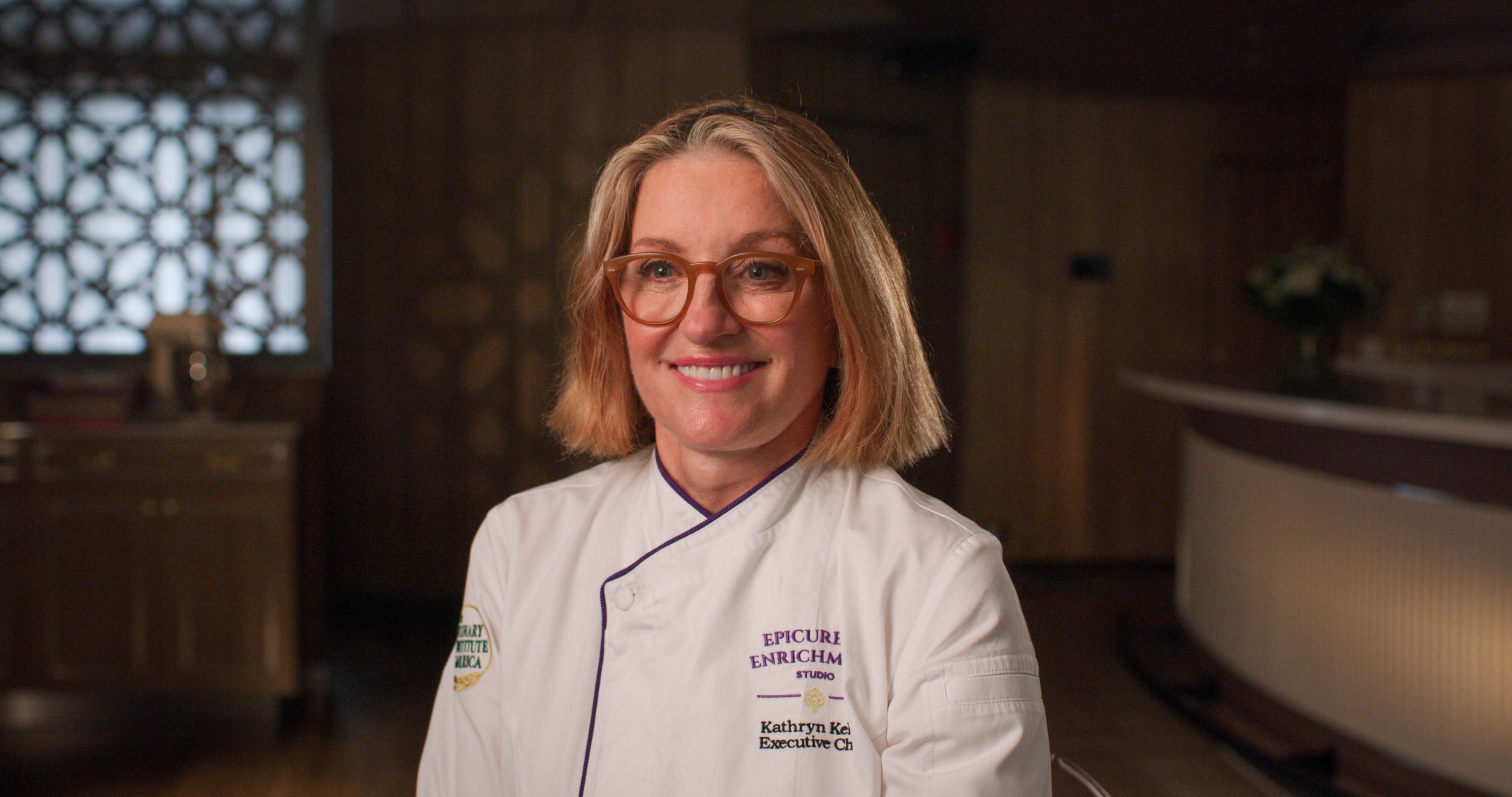 Portrait of a female chef wearing glasses and a white chef's coat, smiling inside a restaurant kitchen with wooden walls and decorative window.
