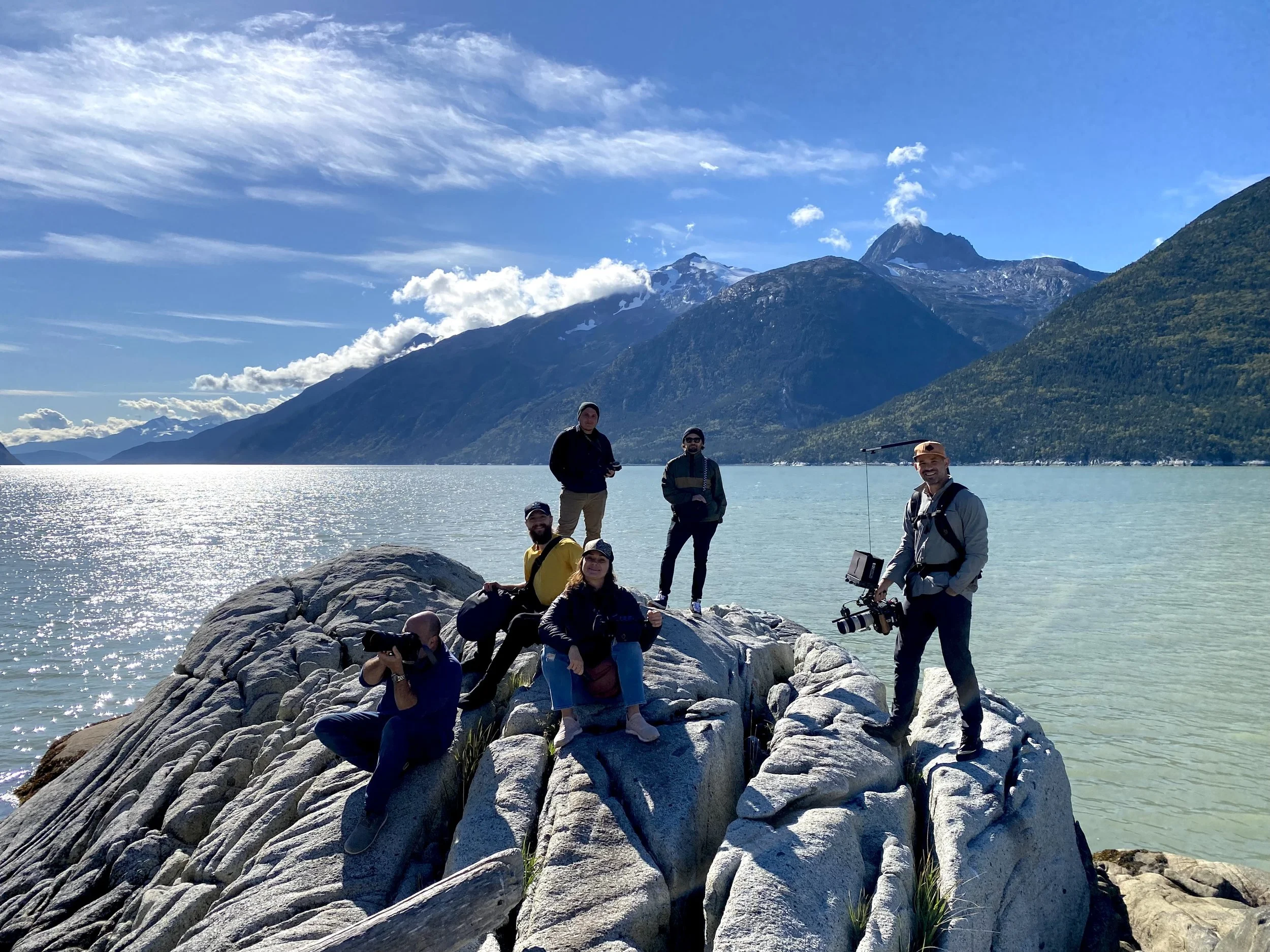 A group of people standing on large rocks near a lake with mountains and a partly cloudy blue sky in the background.