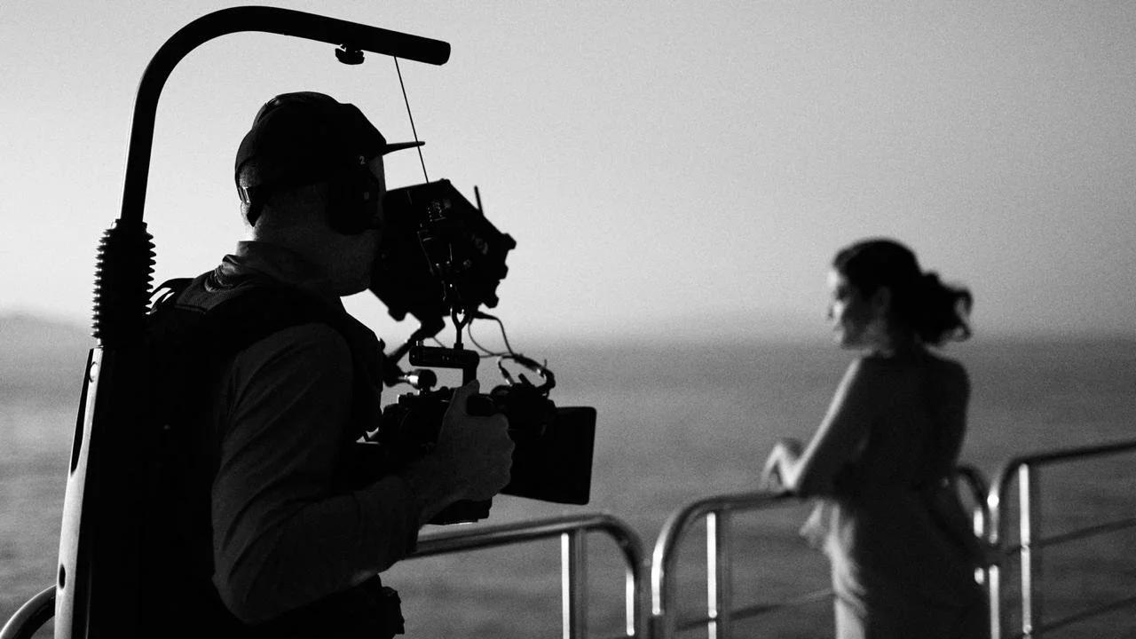 A person operating a professional video camera on a boat or outdoor location, filming a woman standing near the railing with the ocean in the background.