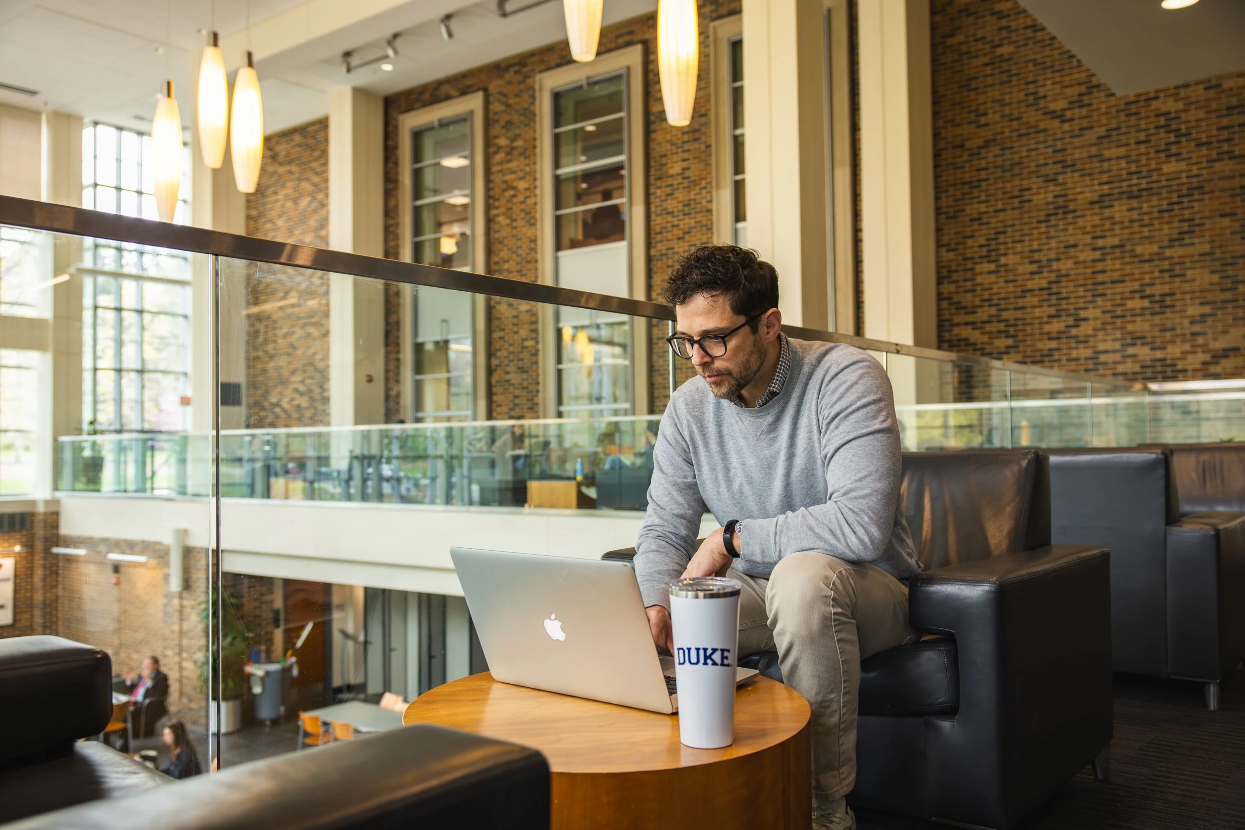 A man with glasses and a beard sitting on a black leather couch, working on a silver MacBook laptop in a spacious modern building. There is a white tumbler with blue text reading 'DUKE' on the wooden table in front of him. The interior features large