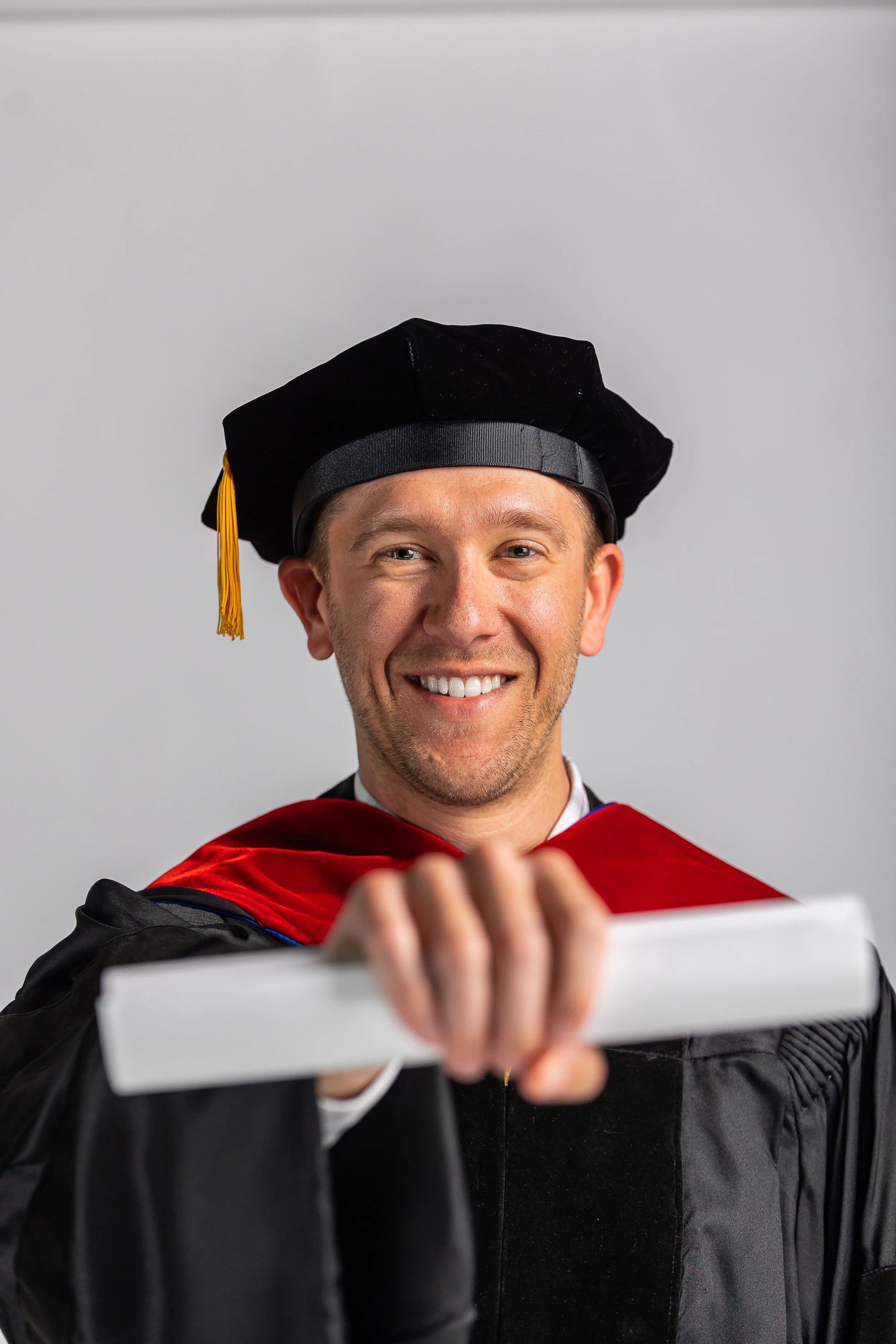 A smiling man in academic regalia taking a selfie with a smartphone. He wears a black graduation cap with a yellow tassel and a red and black gown, standing against a plain gray background.