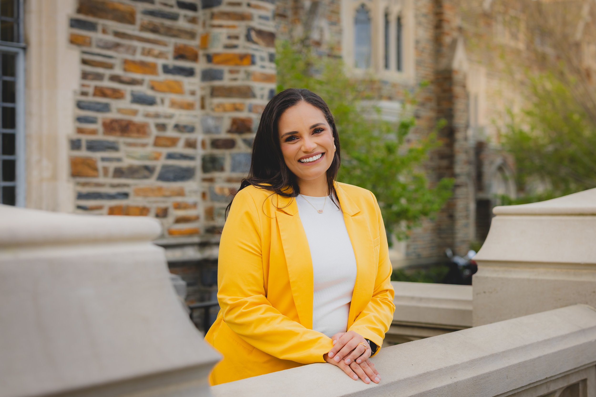 A woman with long dark hair, wearing a white top and yellow blazer, smiling outdoors in front of a stone building with greenery.