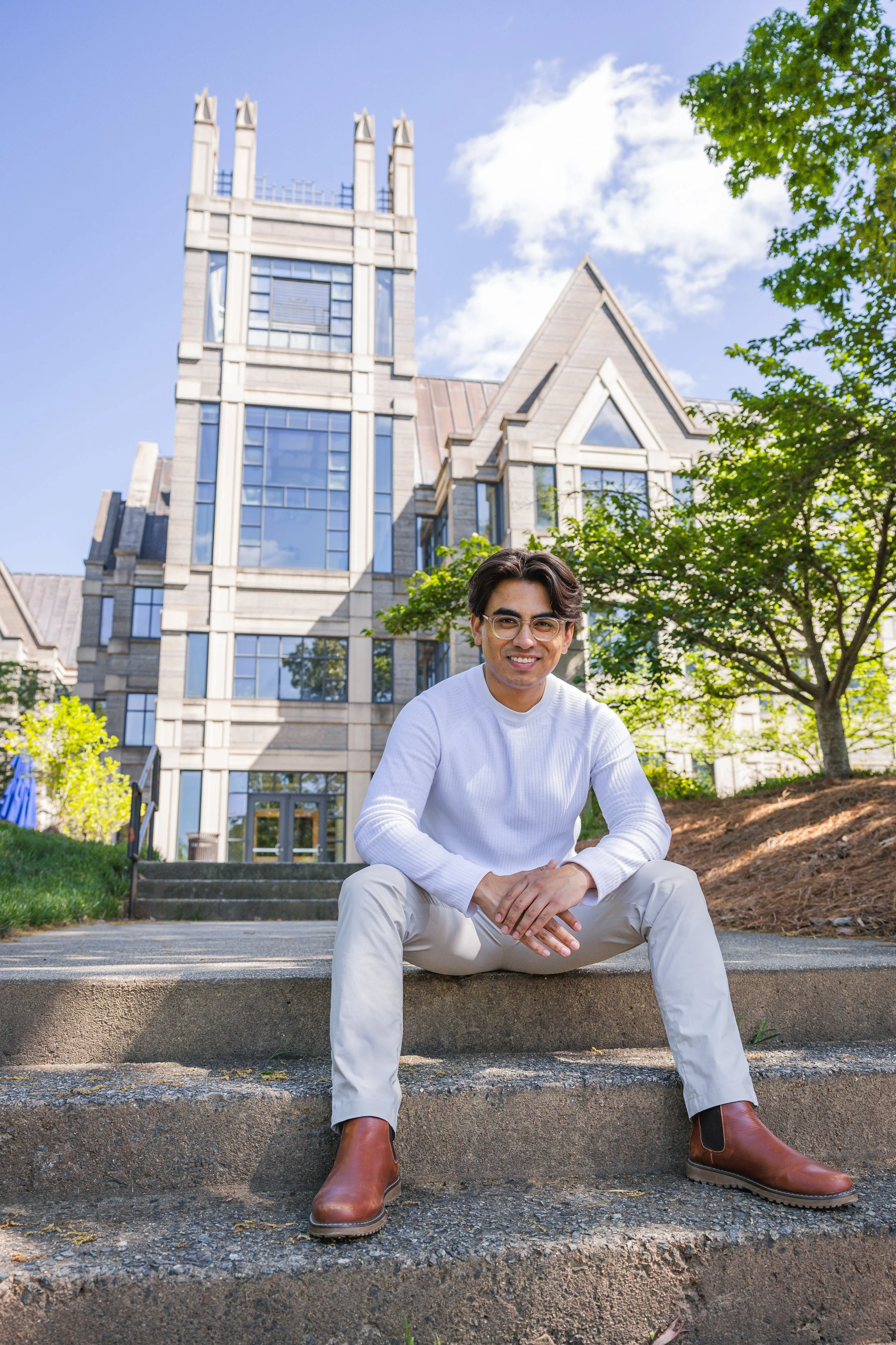 A young man with dark hair, glasses, and a white long-sleeve shirt sitting on outdoor steps with a large modern building and trees in the background.