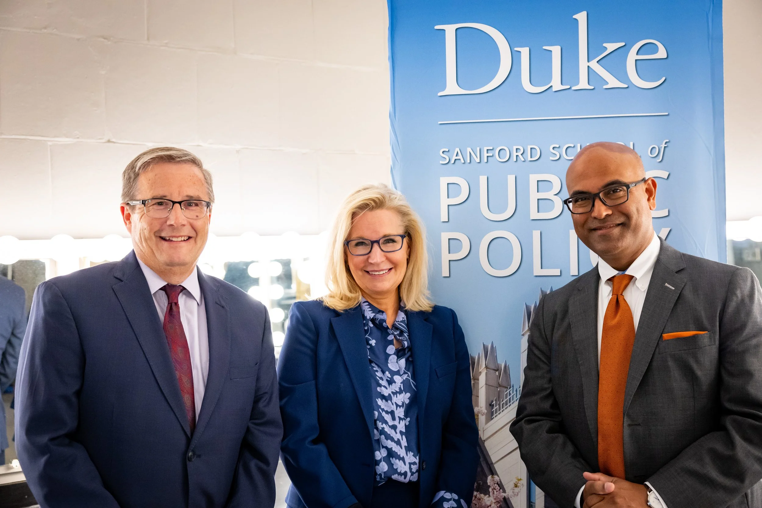 Three professionally dressed individuals standing in front of a blue banner that reads "Duke Sanford School of Public Policy" at an indoor event.
