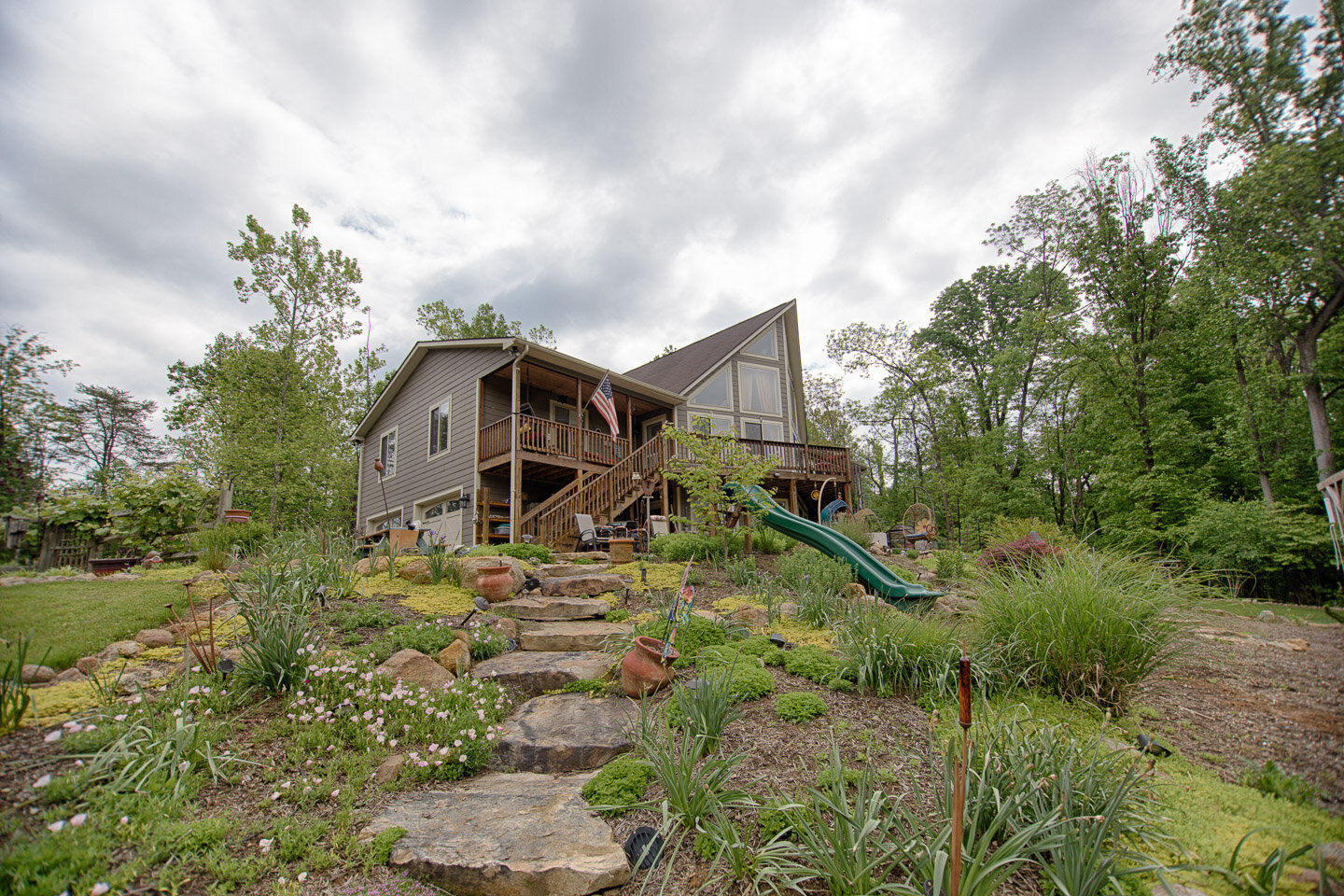 A house with a steep roof and large windows, elevated on stilts with a wooden deck and stairs, surrounded by green trees and a landscaped yard with plants, rocks, and a slide.