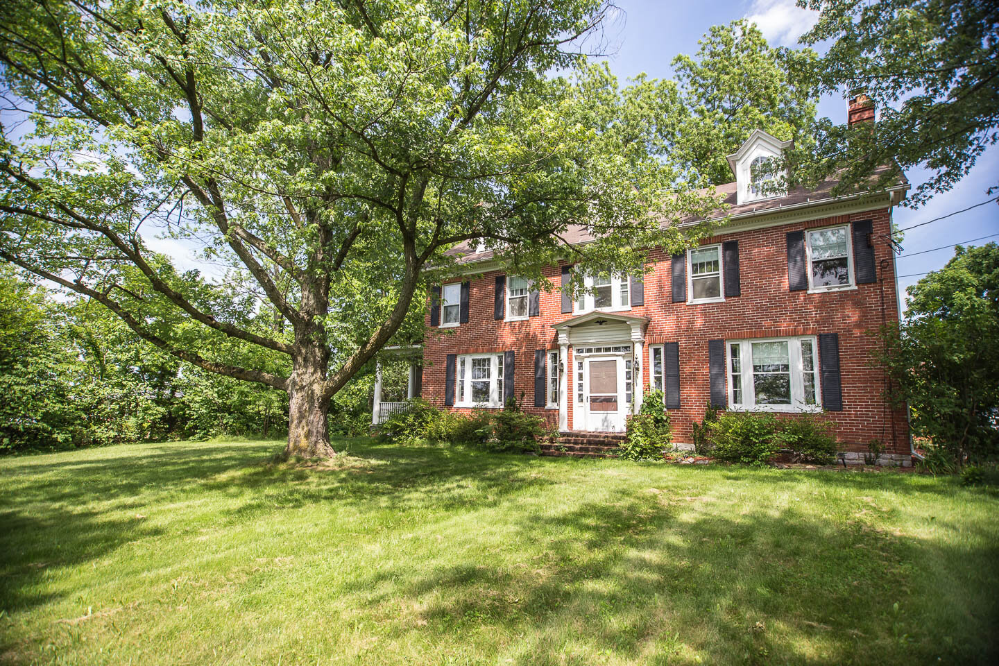 A large brick house with white trim and black shutters, surrounded by green trees and grass on a sunny day.