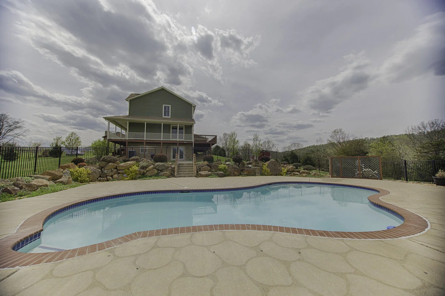 Outdoor view of a swimming pool with a house in the background, a stone wall, plants, and a cloudy sky.