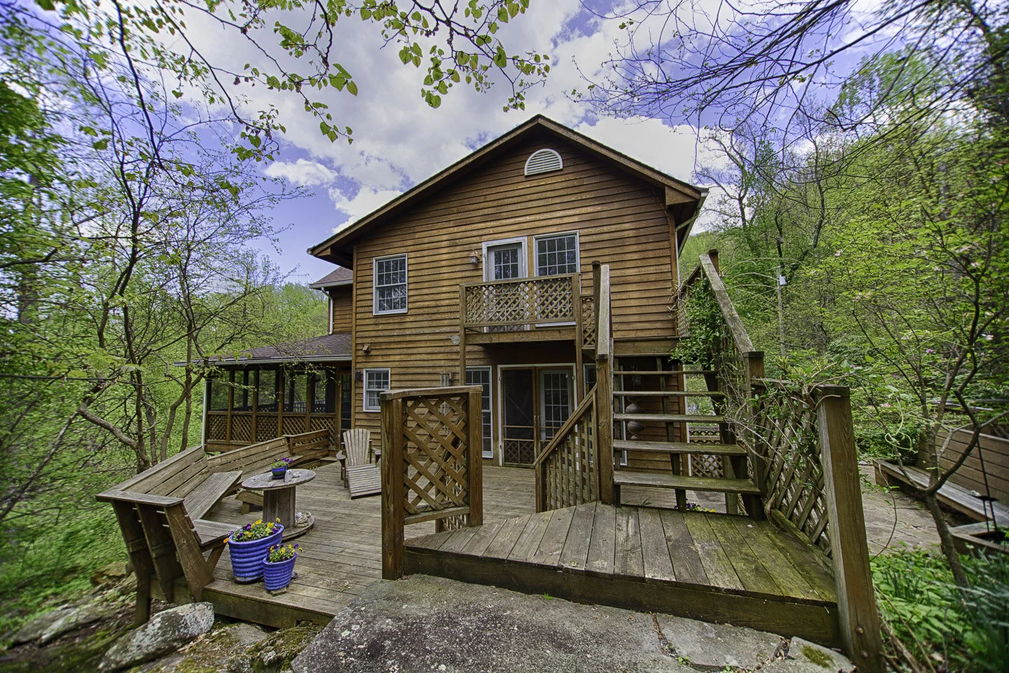 Wooden house with multiple decks and stairs surrounded by trees, with potted plants and outdoor seating.