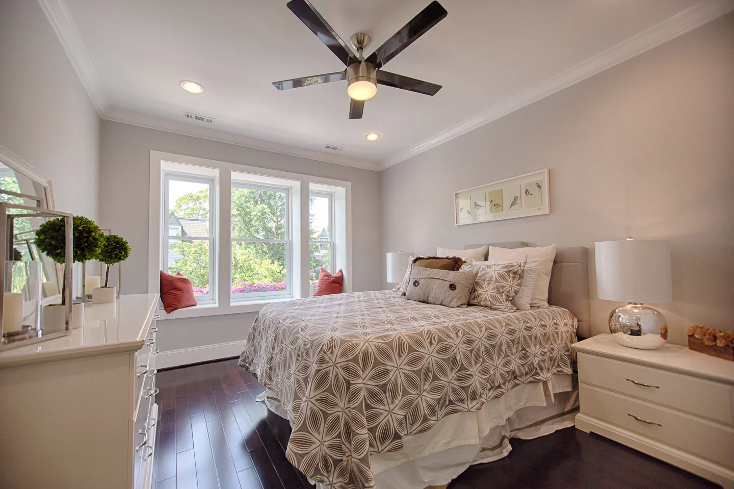A bedroom with a large bed, beige patterned bedspread, multiple pillows, white nightstands with lamps, a window seat with red pillows, and a ceiling fan, with dark hardwood floors and light-colored walls.