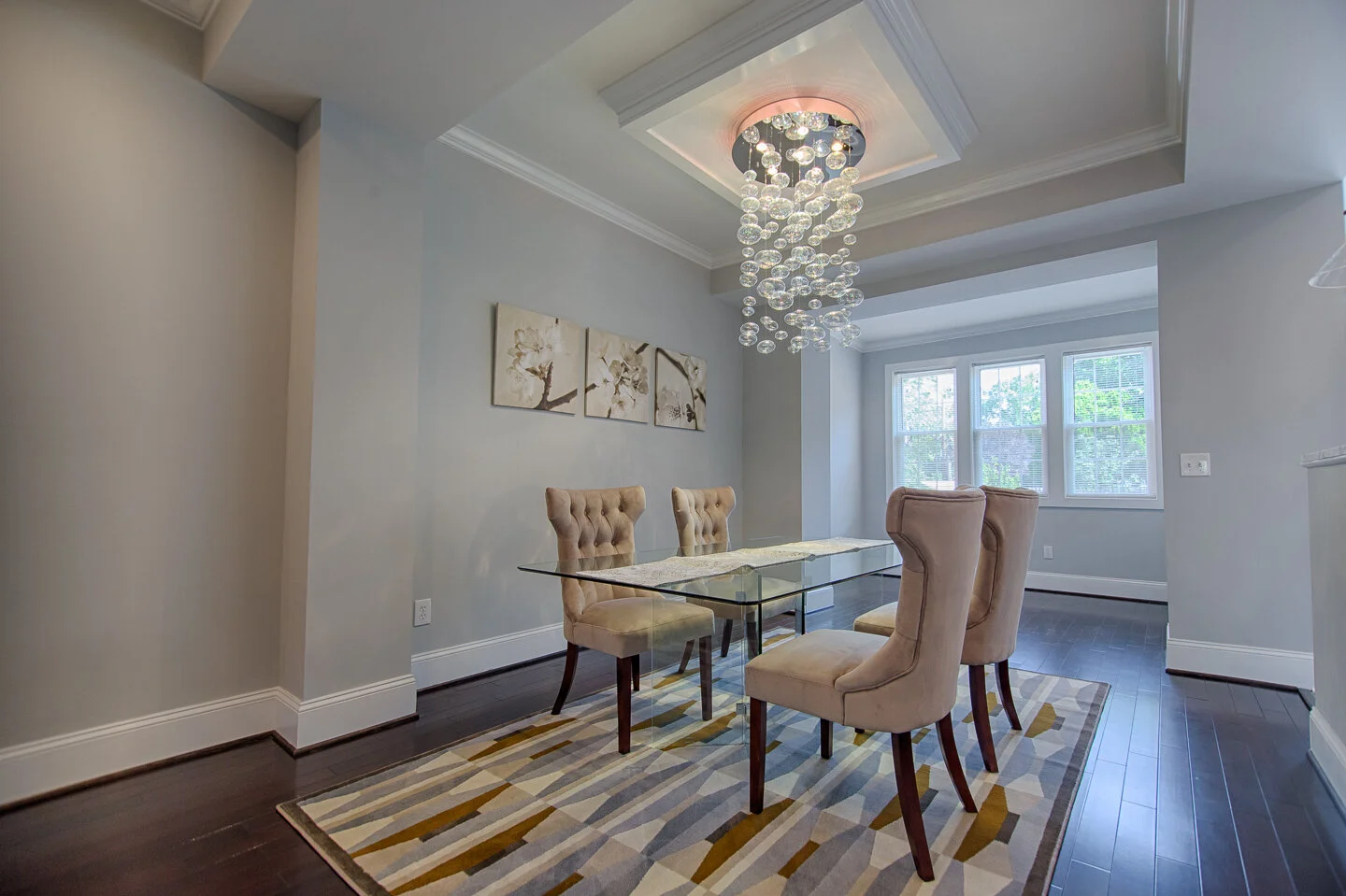 A dining room with a glass table, six beige upholstered chairs, a modern chandelier with hanging glass spheres, a multi-pane window, and a geometric patterned area rug.