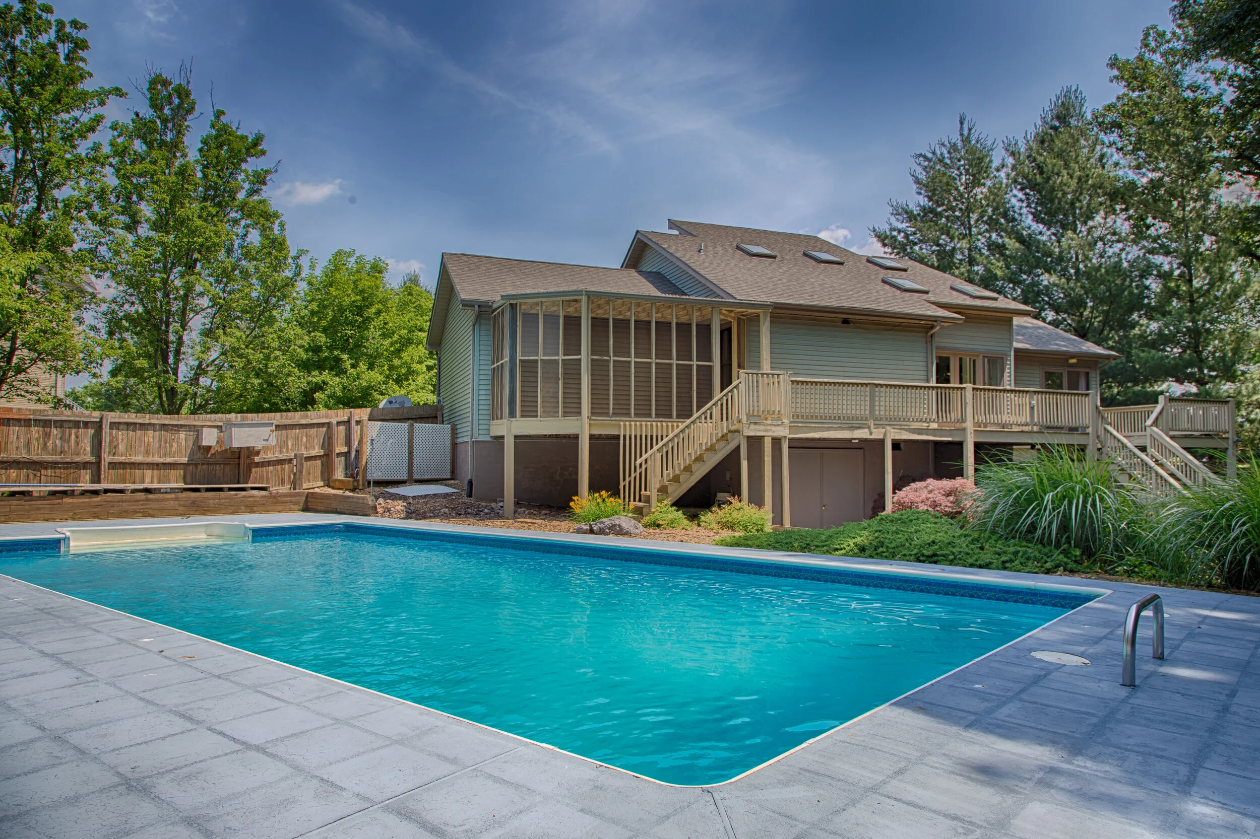 Backyard with a rectangular swimming pool, a wooden deck, and a large house with a screened porch and multiple skylights, surrounded by green trees.