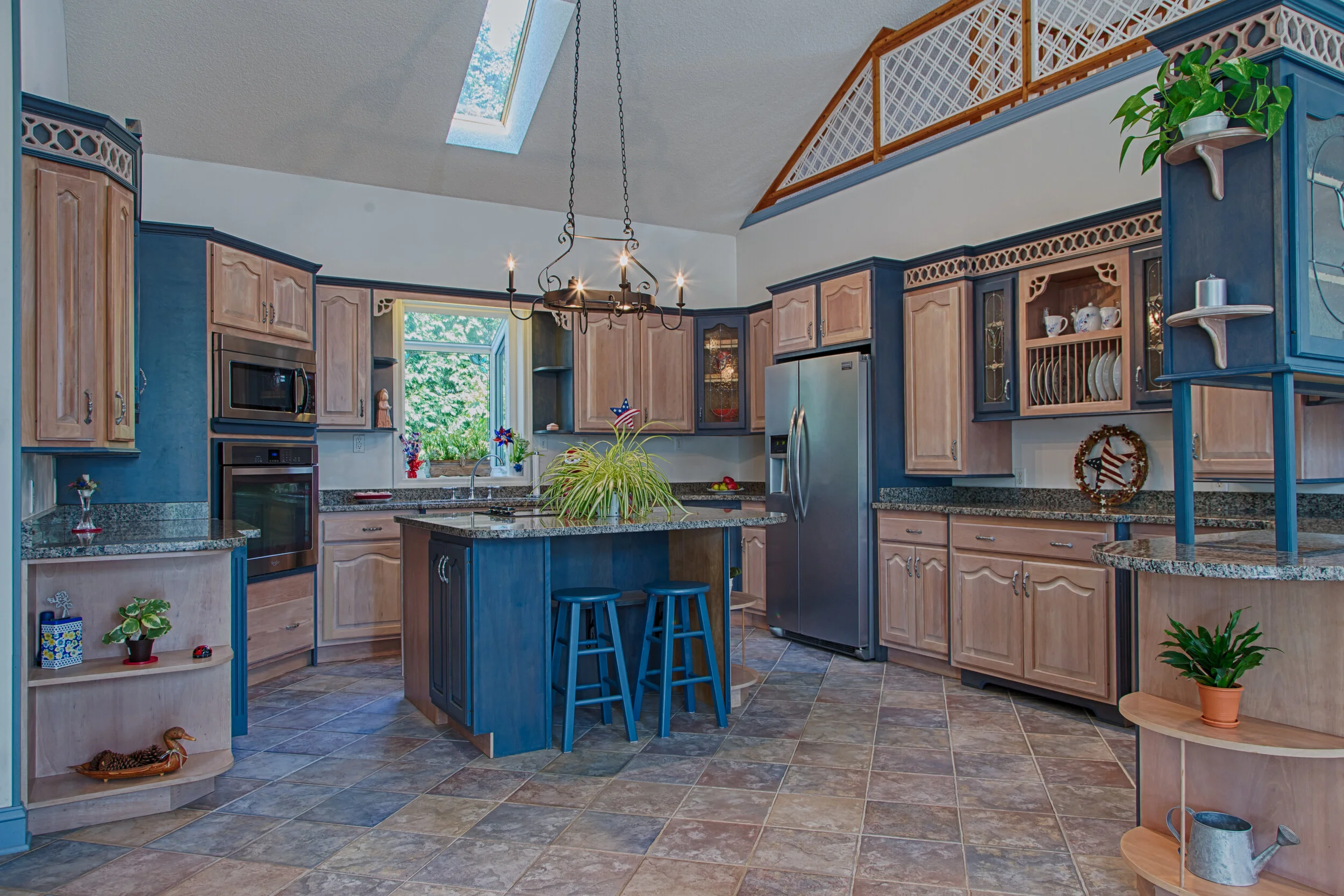 Kitchen with wooden cabinets, granite countertops, a small island with two chairs, stainless steel refrigerator, oven, microwave, and decorative plants and ornaments, with skylight and window letting in natural light.