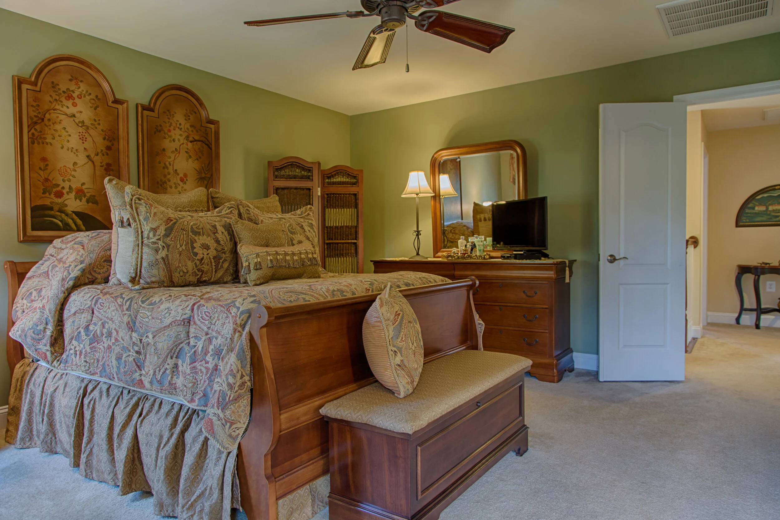 A bedroom with a wooden bed covered in a floral patterned comforter and multiple pillows, a wooden dresser with a mirror and a small television, a green wall, a ceiling fan, and an open door leading to a hallway.