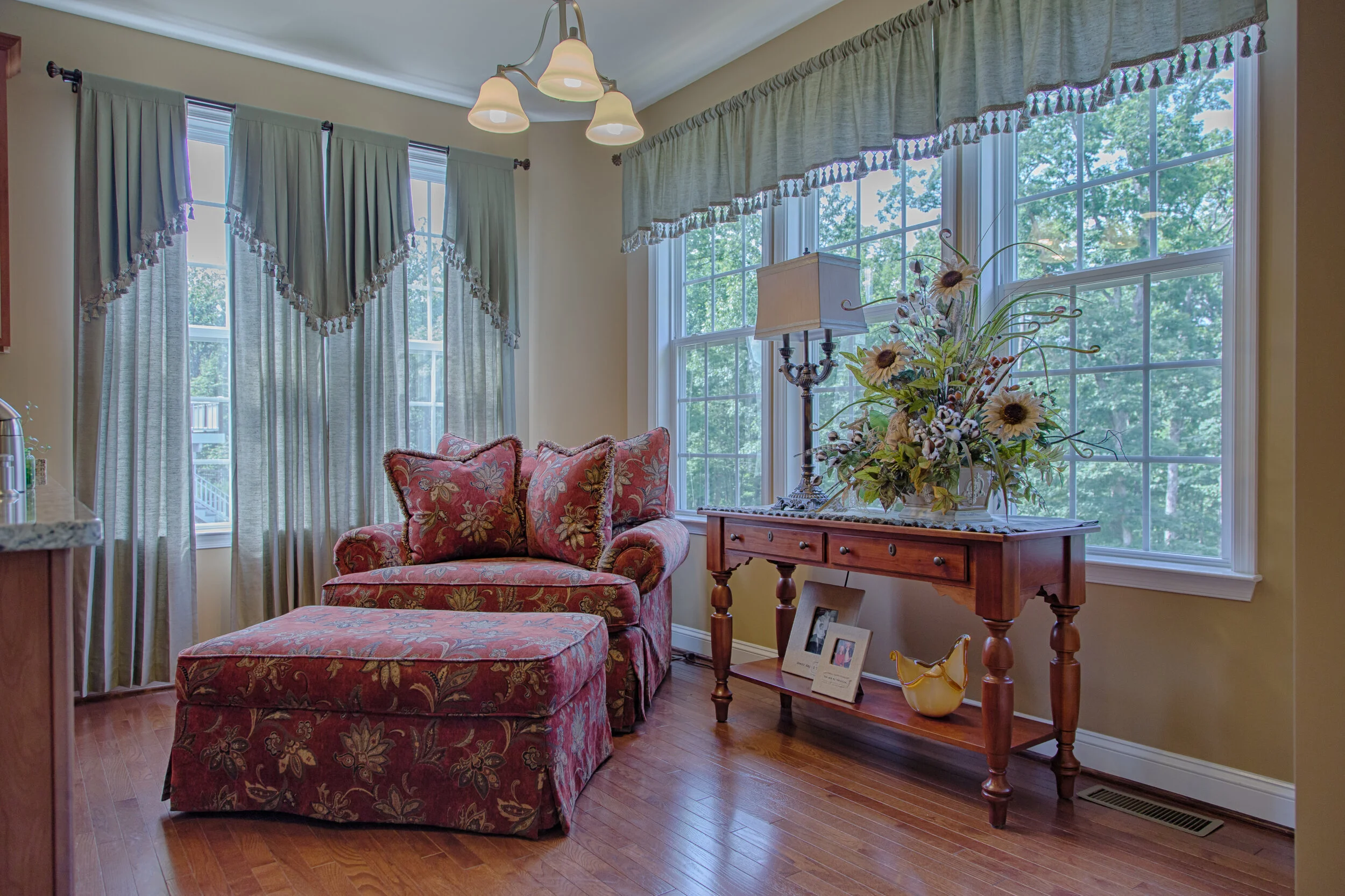 A cozy living room corner with a floral upholstered armchair and ottoman next to a wooden console table holding a floral arrangement, a lamp, and framed photos, in front of large windows with draped curtains.