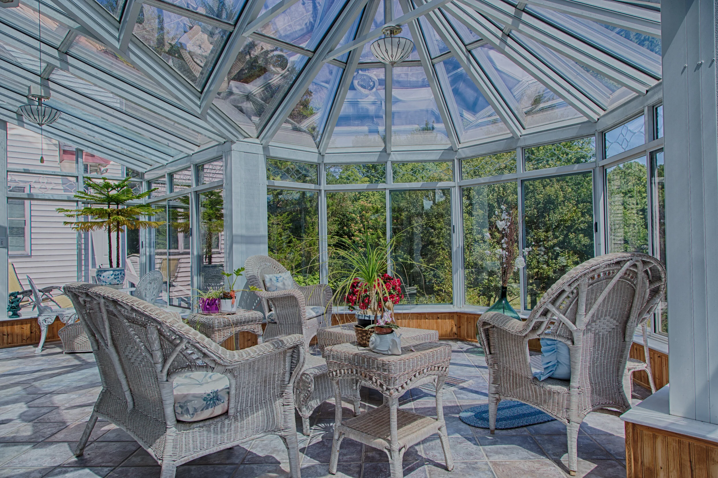Sunroom with wicker chairs, potted plants, and large windows showing green trees outside.