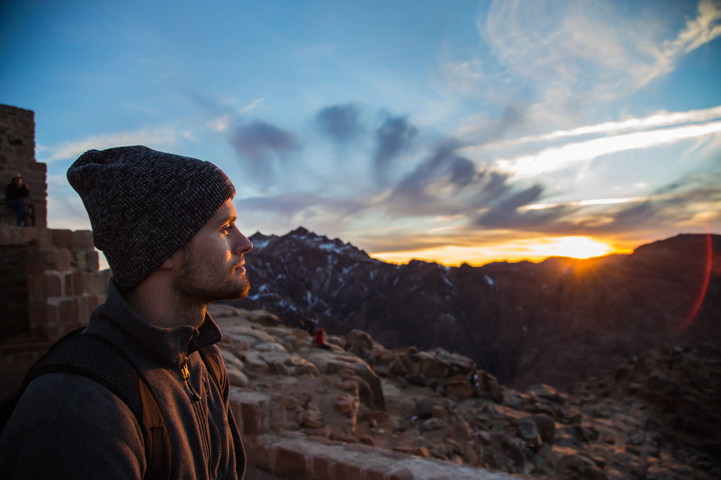 Man wearing a gray beanie and a gray jacket looking at a sunset over mountains with a stone structure nearby.