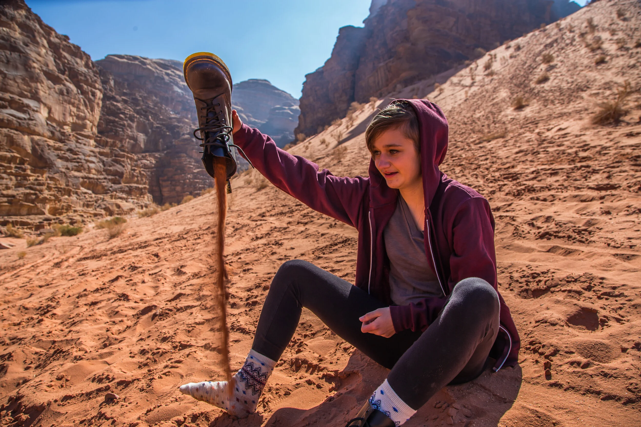 A young woman sitting on sandy ground in a desert landscape, pouring brown liquid from a boot-shaped container, wearing a maroon hoodie, gray shirt, black pants, and patterned socks with mountains in the background.