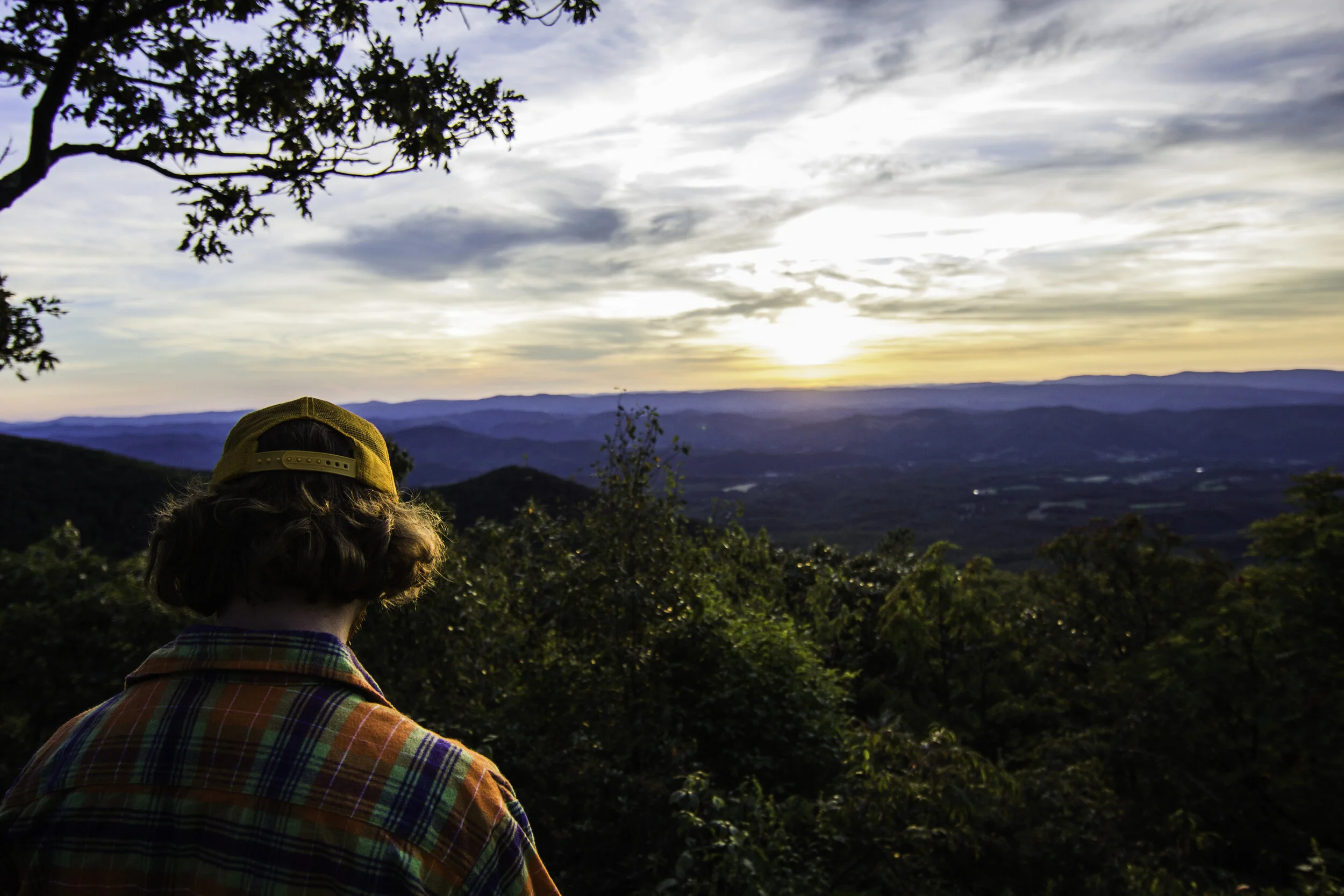 Person with curly hair wearing a yellow cap and plaid shirt looking at a sunset over a mountain landscape with trees in the foreground.