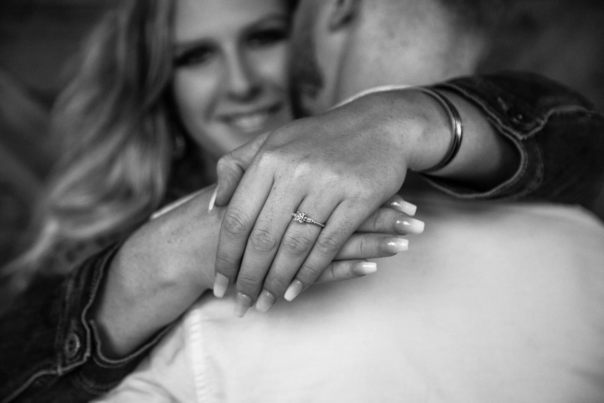 Close-up of a woman’s hand with an engagement ring, resting on another person’s shoulder, smiling in the background.