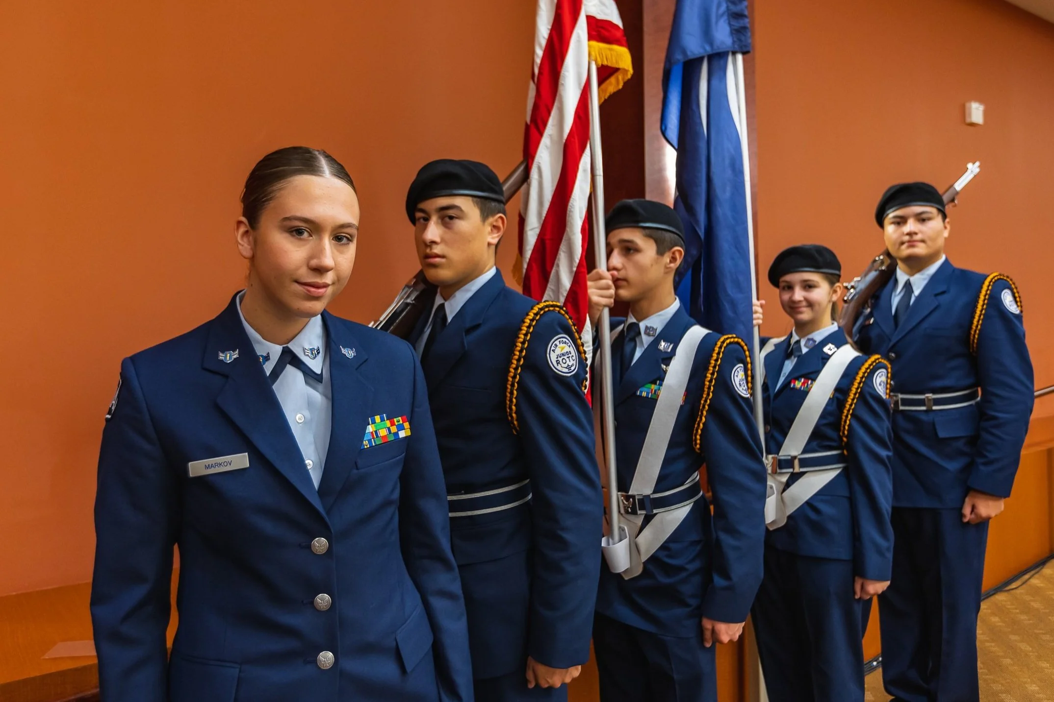 A group of five young cadets in uniform standing in line, holding flags, in a room with an orange wall.