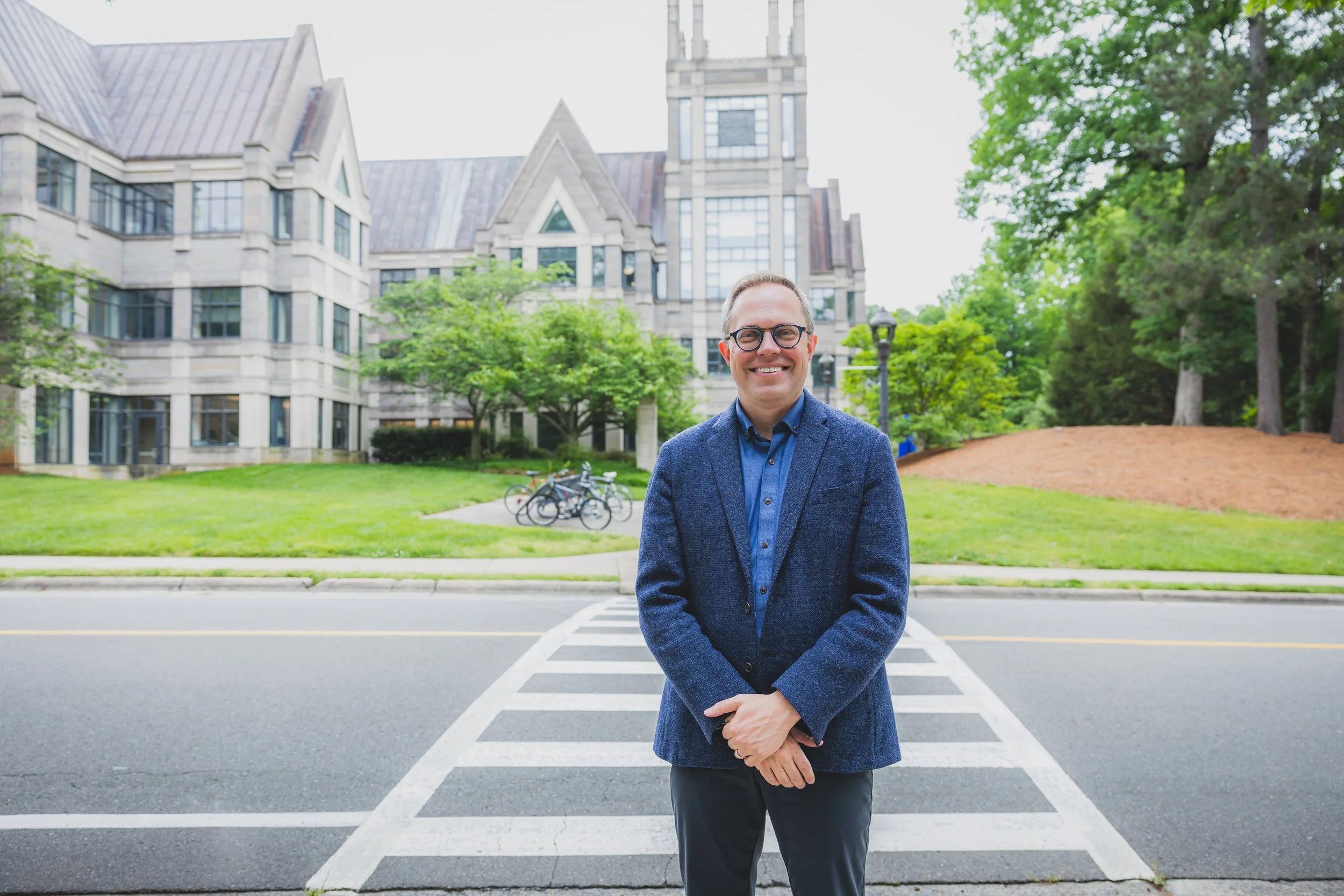 A man standing in front of a crosswalk on a city street, smiling, with a large historic building, trees, and bicycles in the background.